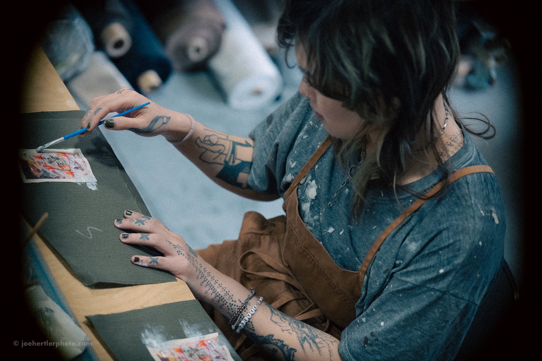 A woman with tattoos and dark hair painting on a small canvas at a desk, with spools of thread in the background.