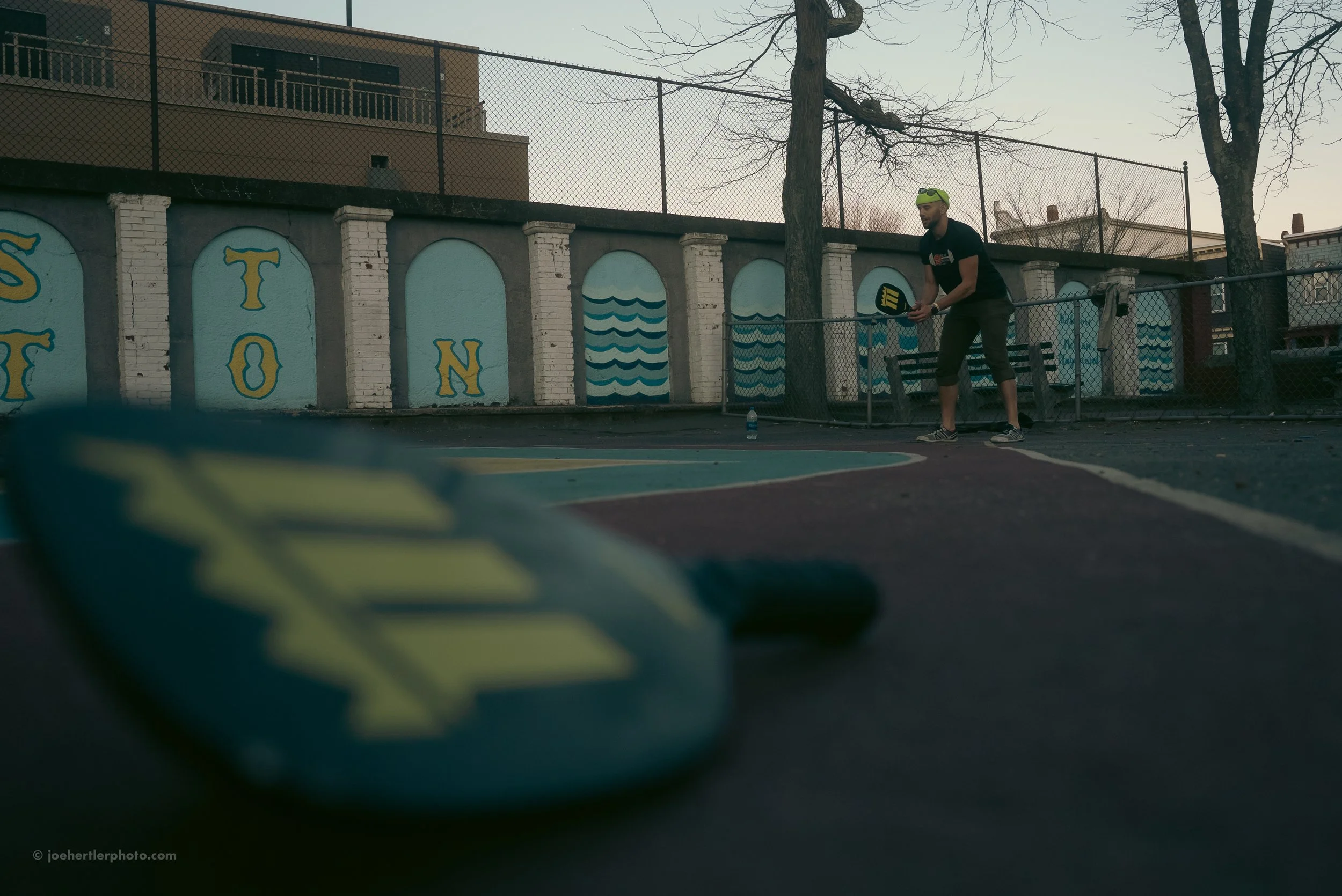 Person practicing pickleball on an outdoor court with mural art wall in the background, tennis paddle in hand, water bottle on court, pickleball shoe in foreground.