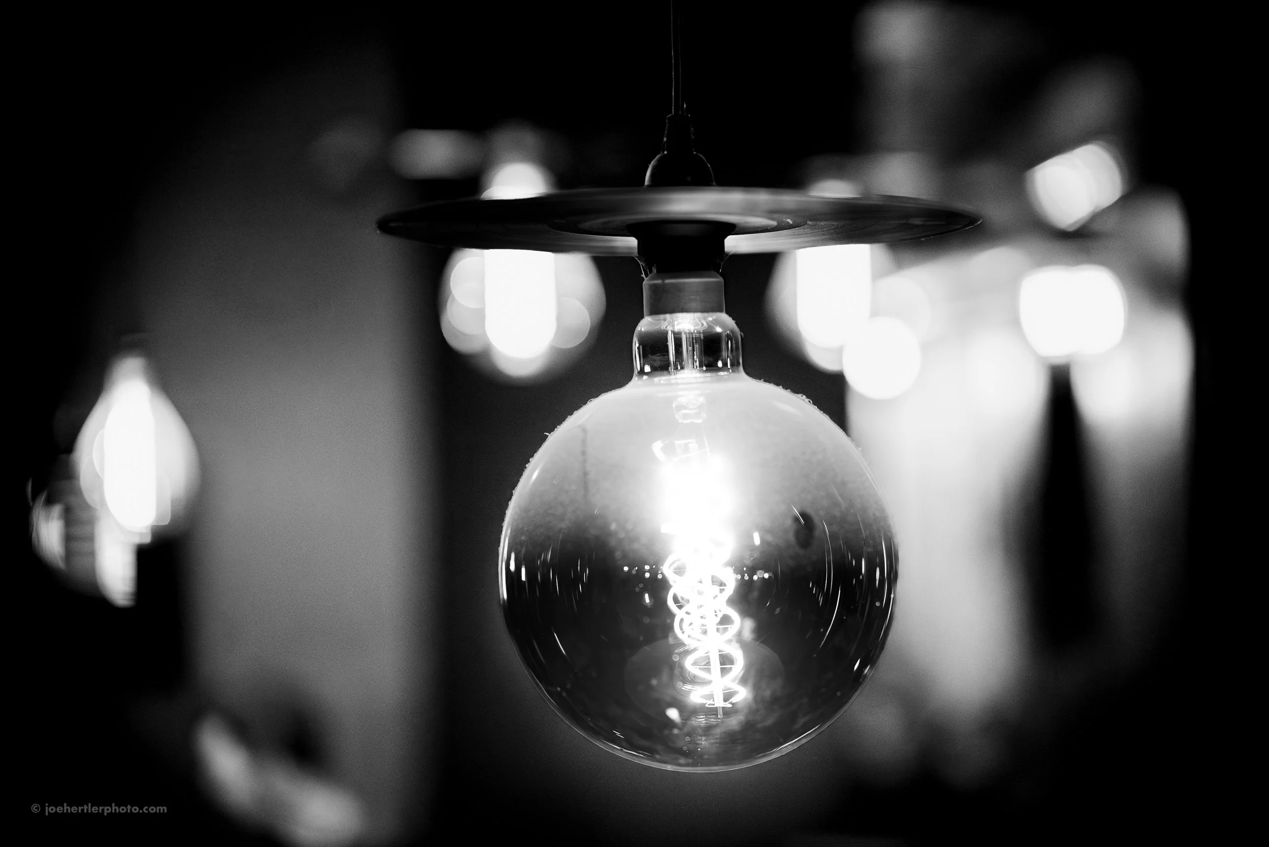 Close-up of an illuminated incandescent light bulb hanging from the ceiling, with a dark background and blurred lights in the distance.