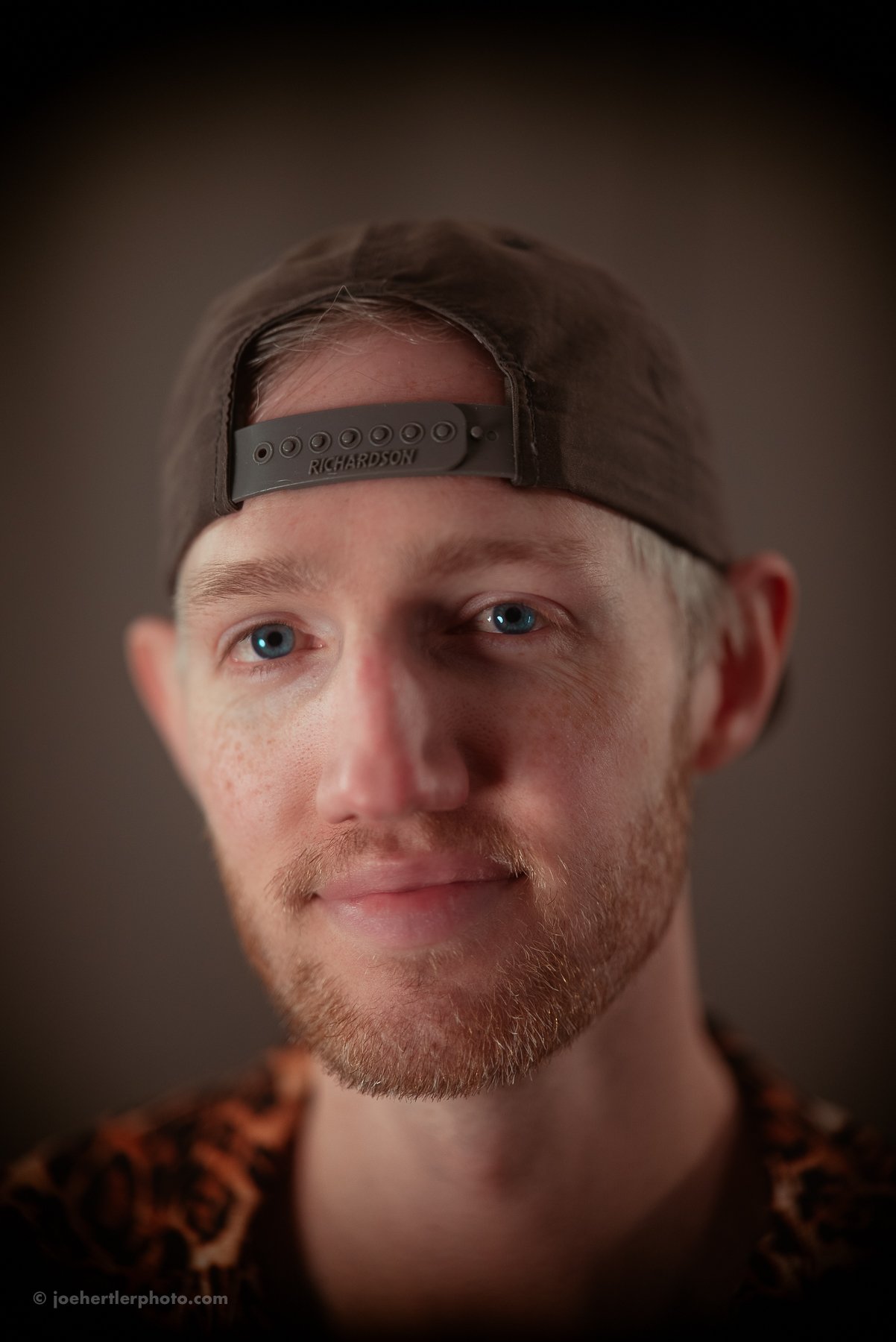 A young man with blond hair and a beard wearing a backwards cap and a leopard print shirt, looking slightly to the side.