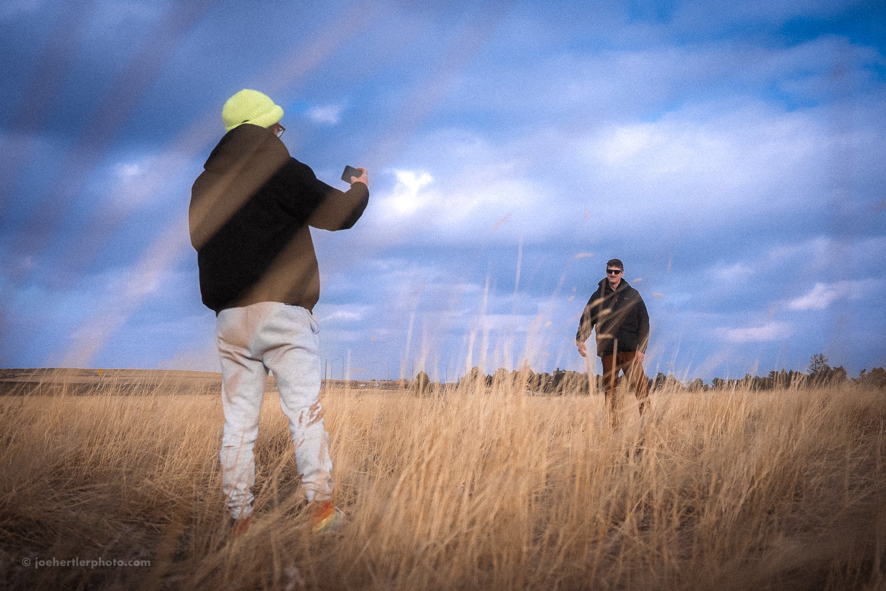 Two men in a grassy field under a cloudy sky; one is taking a photo with a smartphone, the other standing at a distance wearing sunglasses.