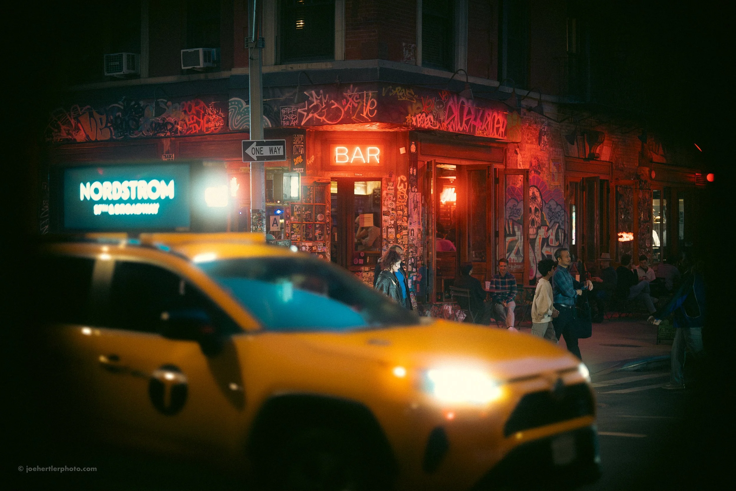Nighttime street scene outside a bar with neon signs, graffiti-covered walls, people sitting and walking, and a yellow taxi cab in the foreground.