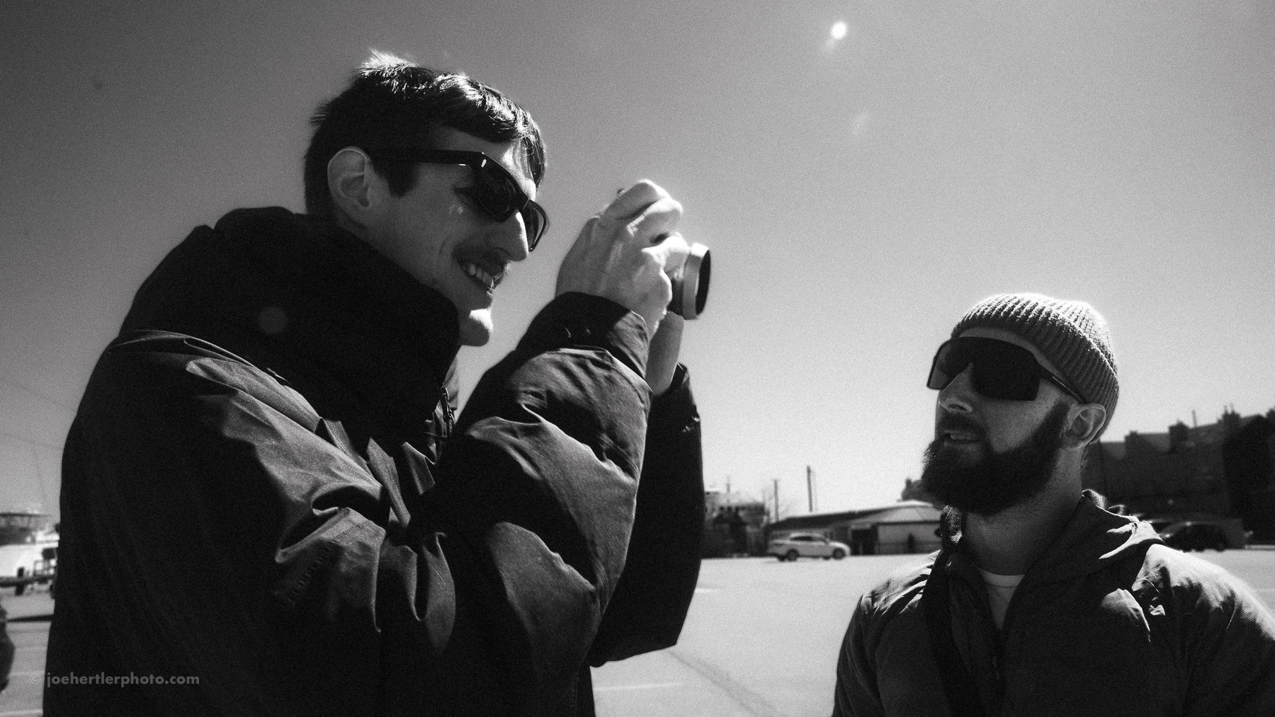 Two men outdoors, one taking a photo with a small camera, both wearing sunglasses and jackets, in a bright environment with cars and buildings in the background.