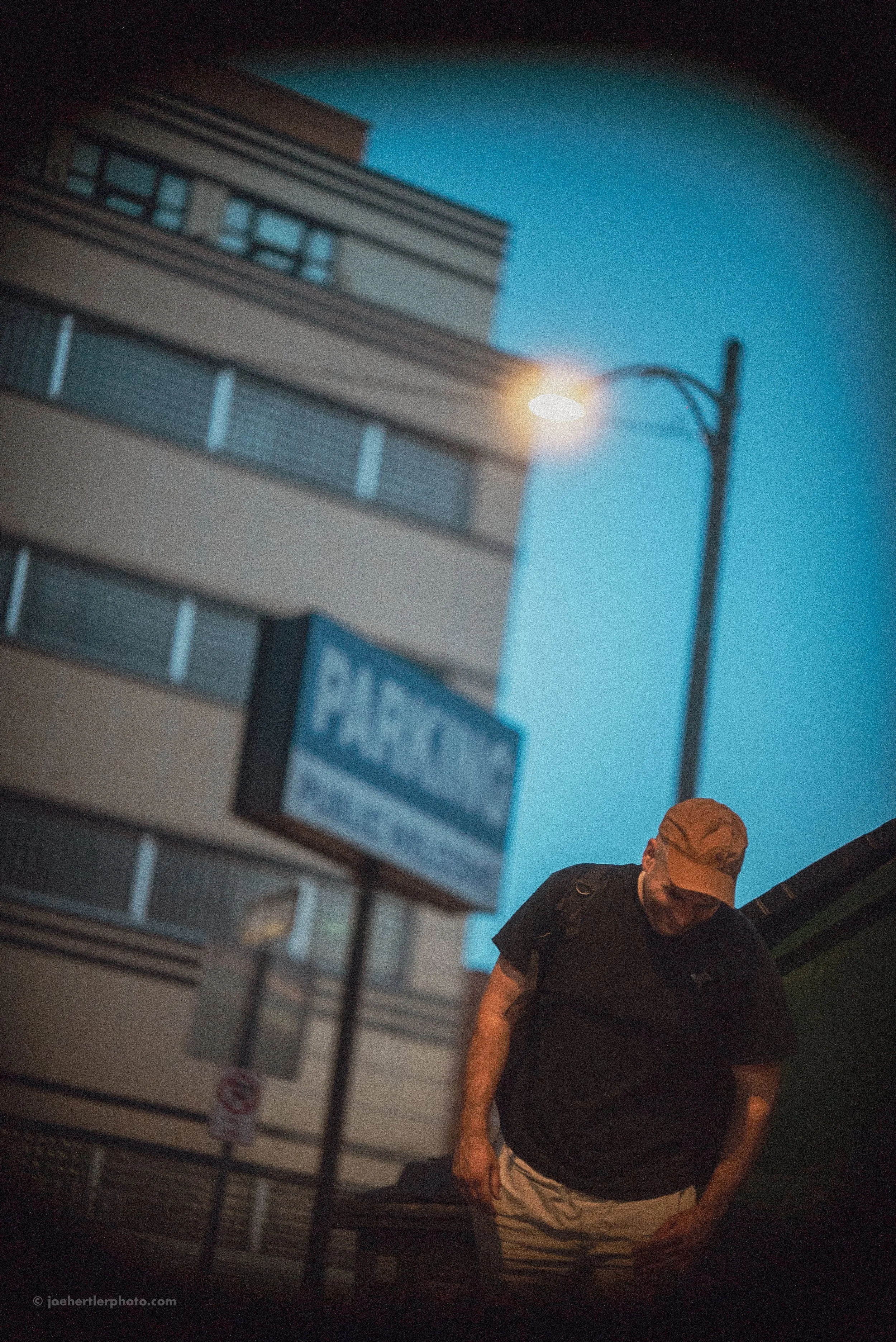 A man in a black t-shirt and beige cap leaning against a green object outdoors at dusk with a multi-story building, streetlamp, and parking sign in the background.