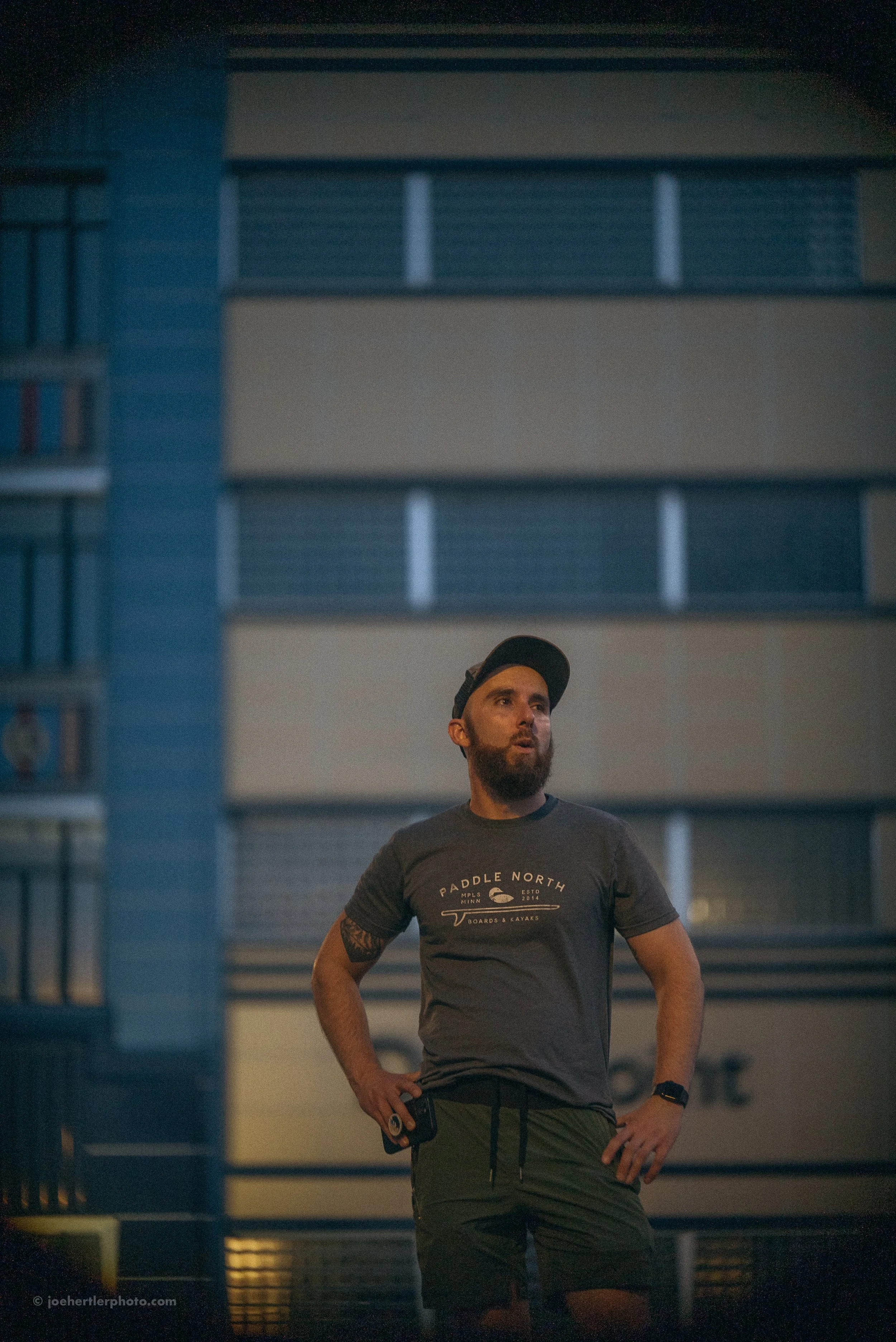 A man with a beard and tattoos wearing a black cap, gray t-shirt with a Paddle North logo, and dark shorts, stands outdoors at night with hands on hips, looking to the side.