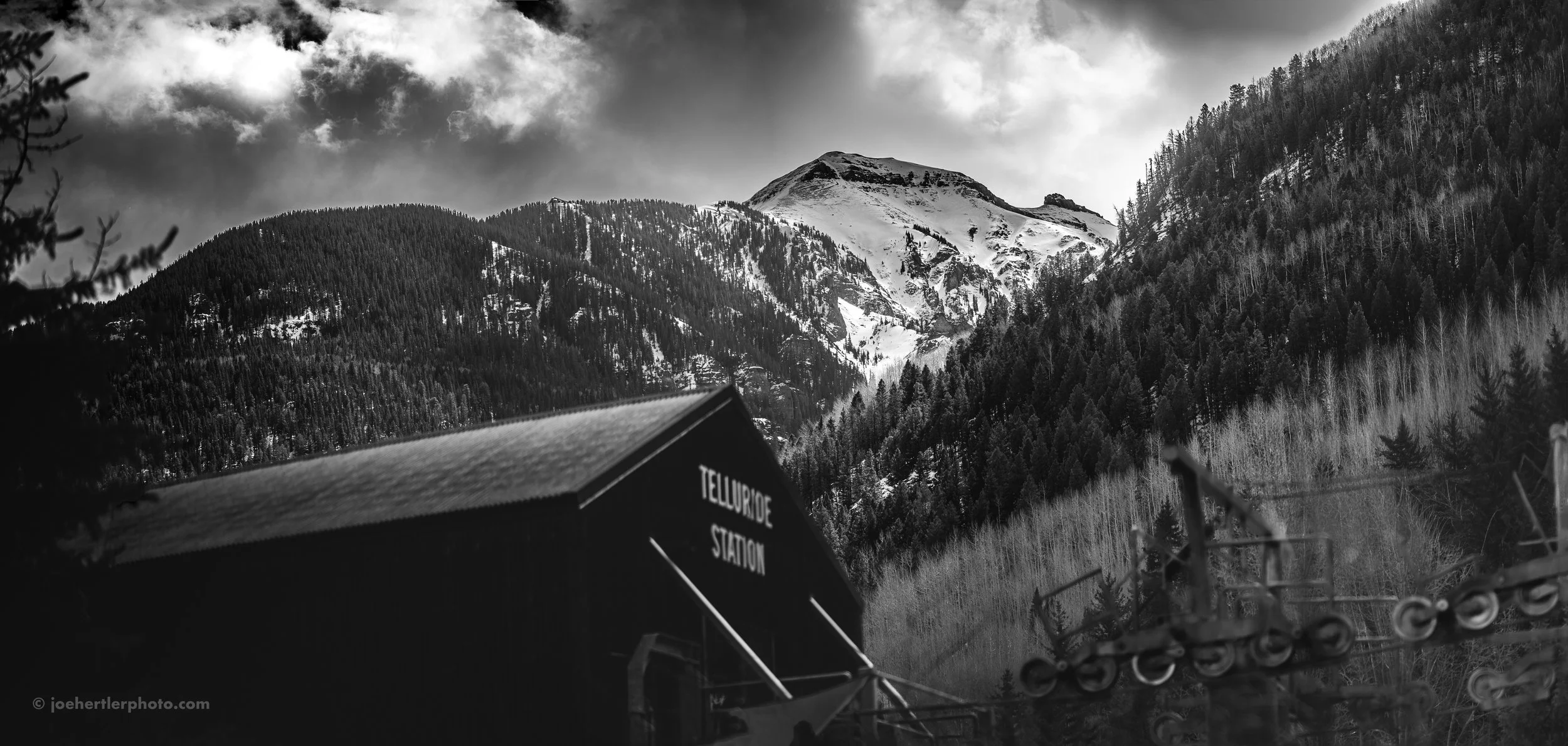 Black and white photo of snowy mountain landscape with a ski lift station labeled 'Telluride Station' in the foreground.