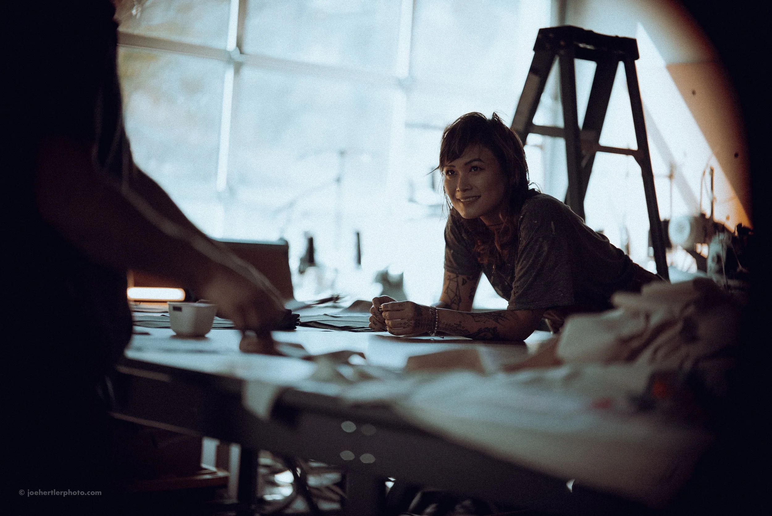 A woman with tattoos smiling and leaning on a table in a workshop, with a person arranging papers in the foreground and a ladder in the background.