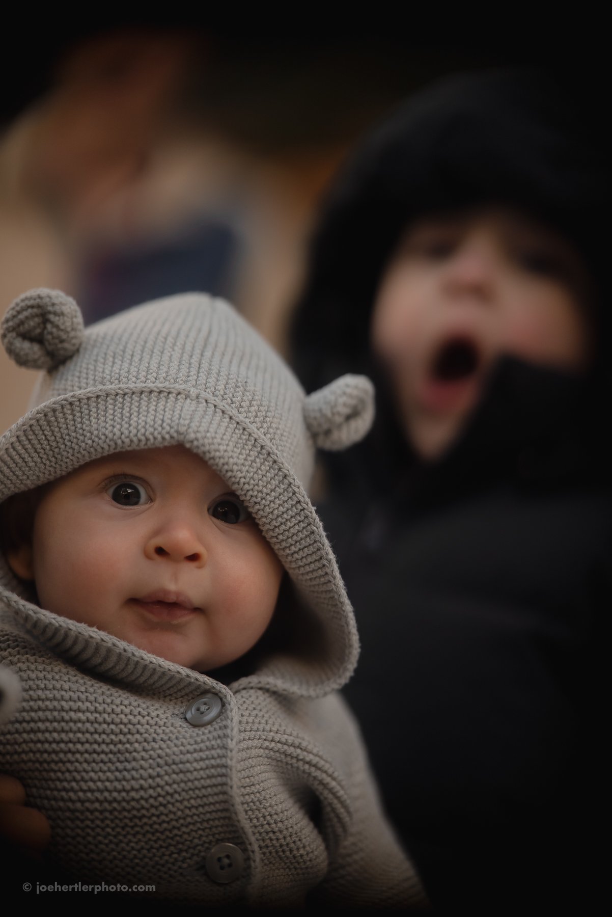 Close-up of a baby wearing a beige hooded sweater with small bear ears, looking directly at the camera. Behind, a child with an open mouth, wearing a black hooded jacket, appears to be excited or surprised, with other blurred figures in the backgroun