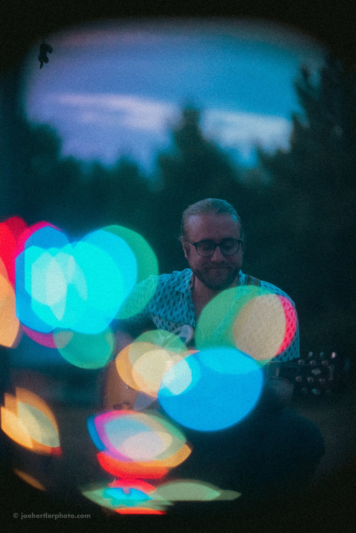 A man with glasses playing guitar outdoors during twilight, with colorful bokeh lights in the foreground and a dark, forested background.