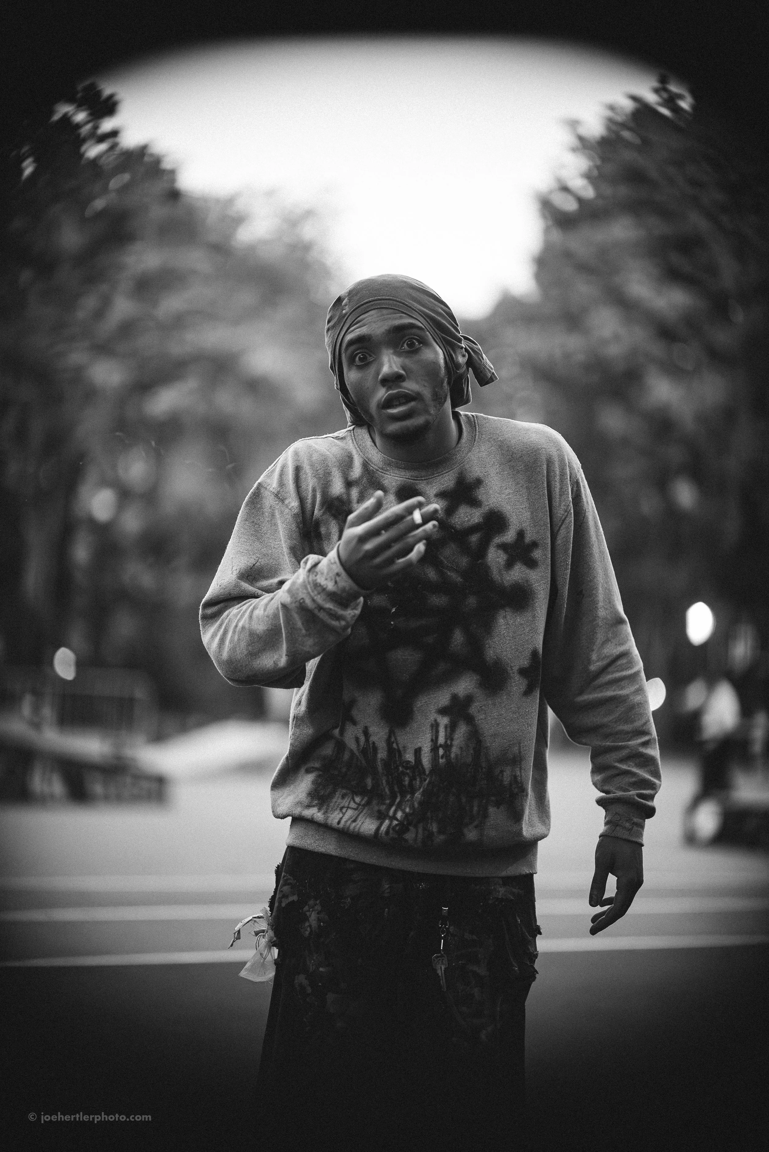 A young man with a bandana on his head, wearing a sweatshirt with spray-painted stars and a city skyline, appears to be in a state of distress or confusion on a street with trees and blurred vehicles in the background.