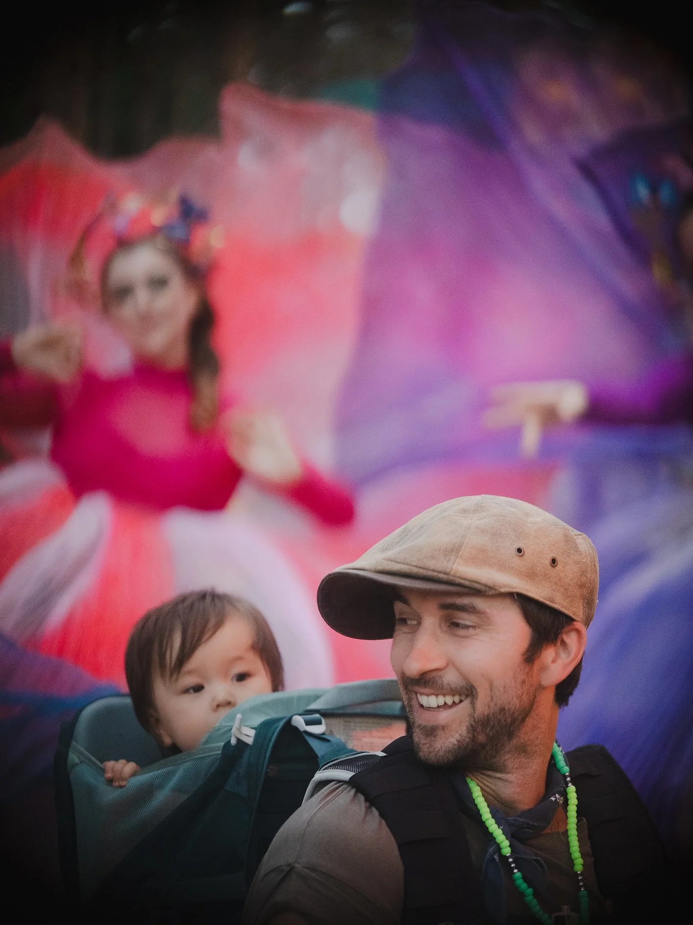 A man smiling with a young child in a backpack carrier, with a animated character princess in the background.