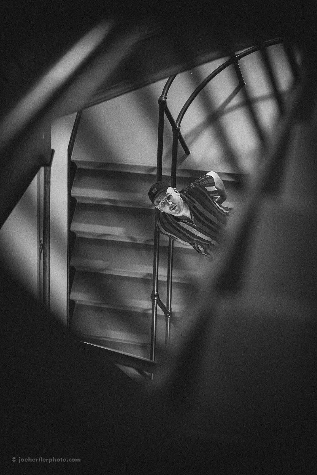 A man is looking up at the camera from a staircase, viewed through a framing of dark objects. The photo is in black and white.
