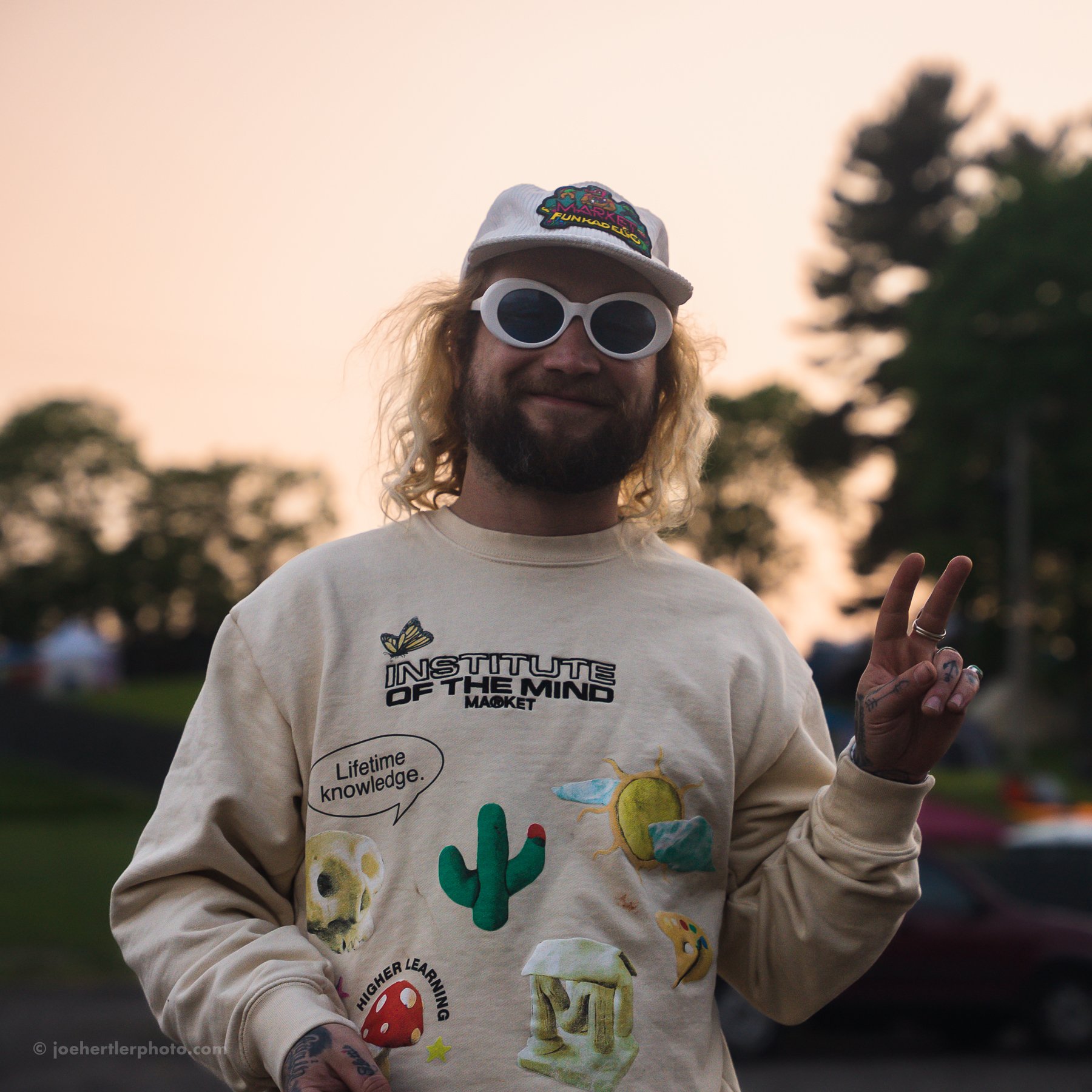 A man with curly blonde hair, beard, wearing white sunglasses, a white cap with a colorful patch, and a beige sweatshirt with various colorful graphics and text, smiling and making a peace sign with his right hand outdoors at sunset.