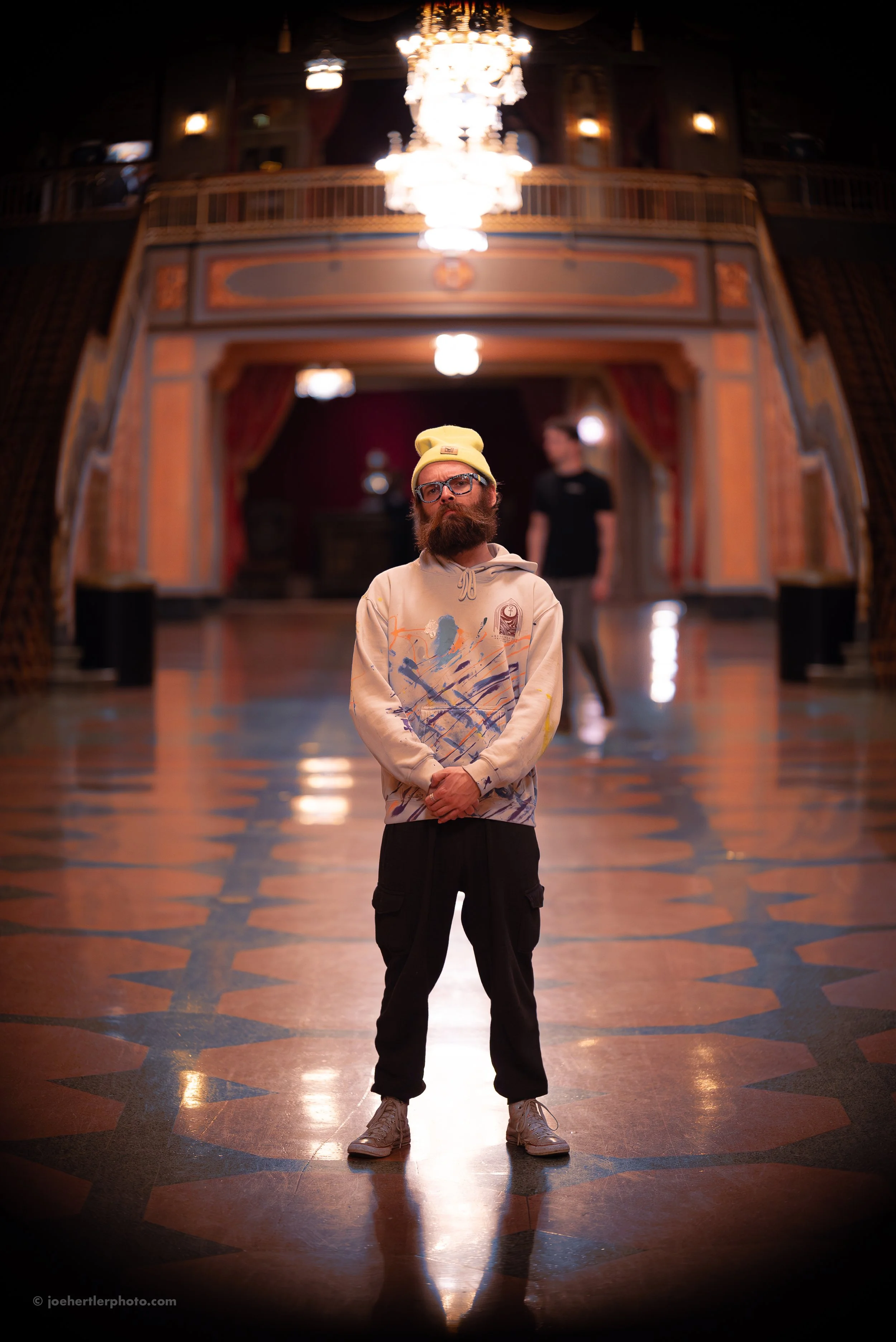 A man wearing a yellow beanie, glasses, a beige hoodie with colorful paint splashes, black cargo pants, and gray sneakers standing in a grand, ornate hall with a chandelier overhead and an elaborate wooden balcony. Another person is blurred in the ba