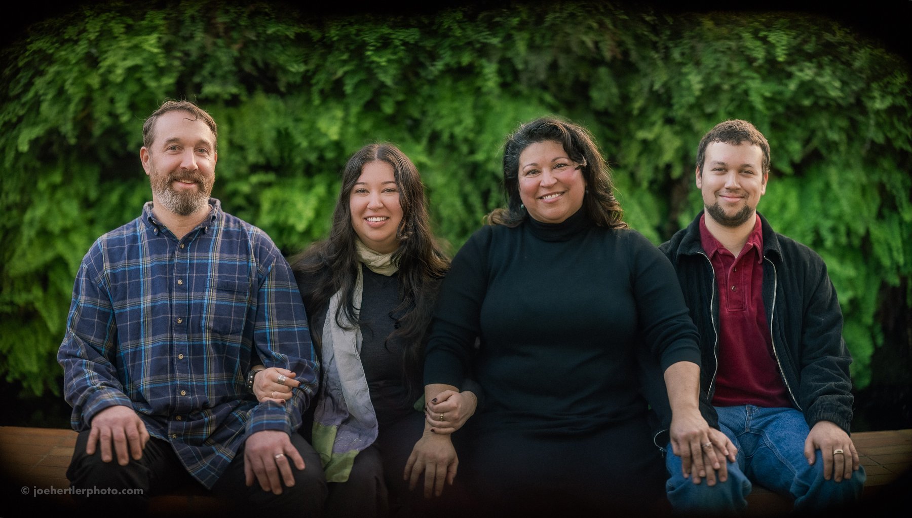 Four people sitting outdoors on a wooden bench with green foliage background, smiling at the camera.