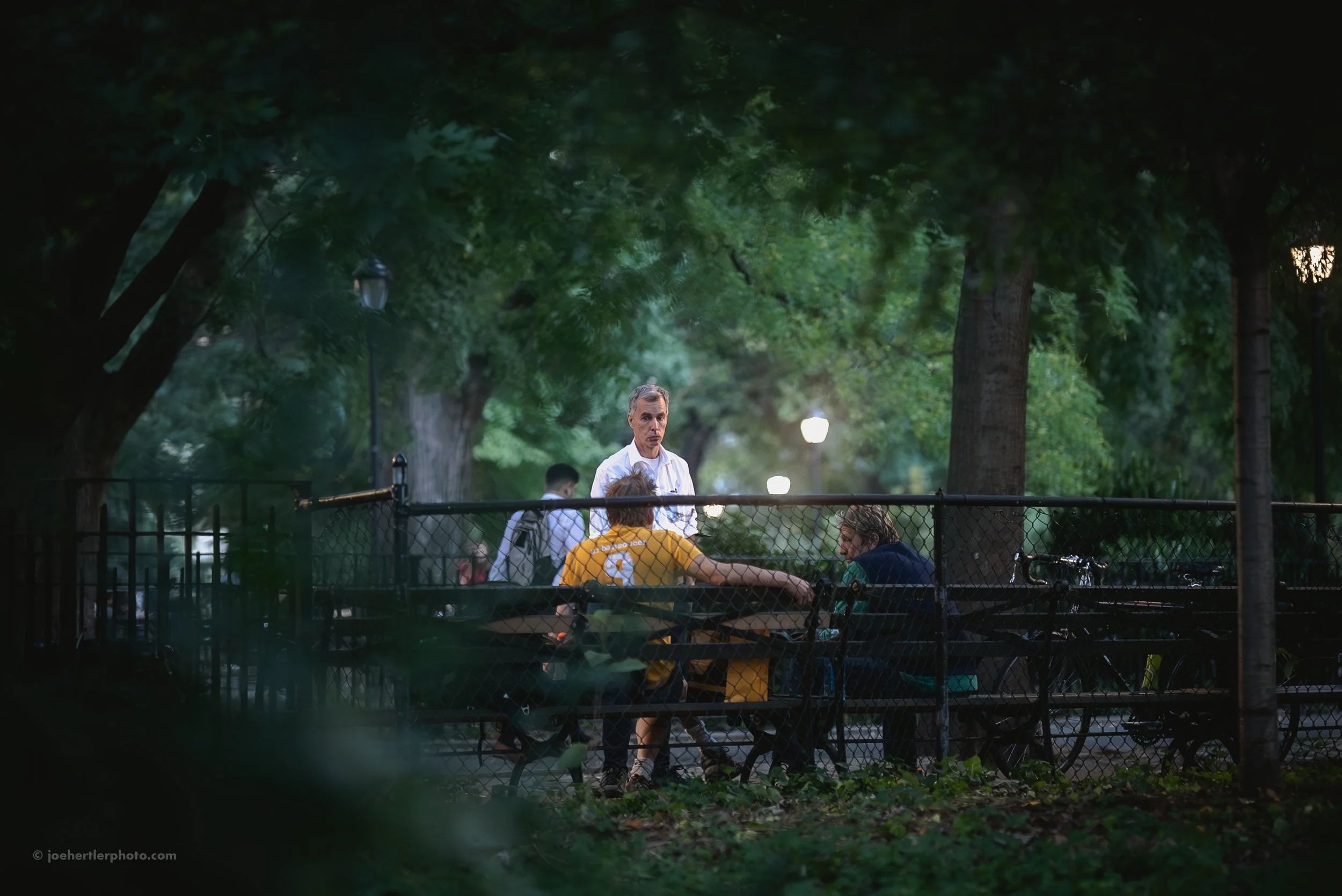 People sitting on benches and talking in a park with trees and streetlights in the background during evening.