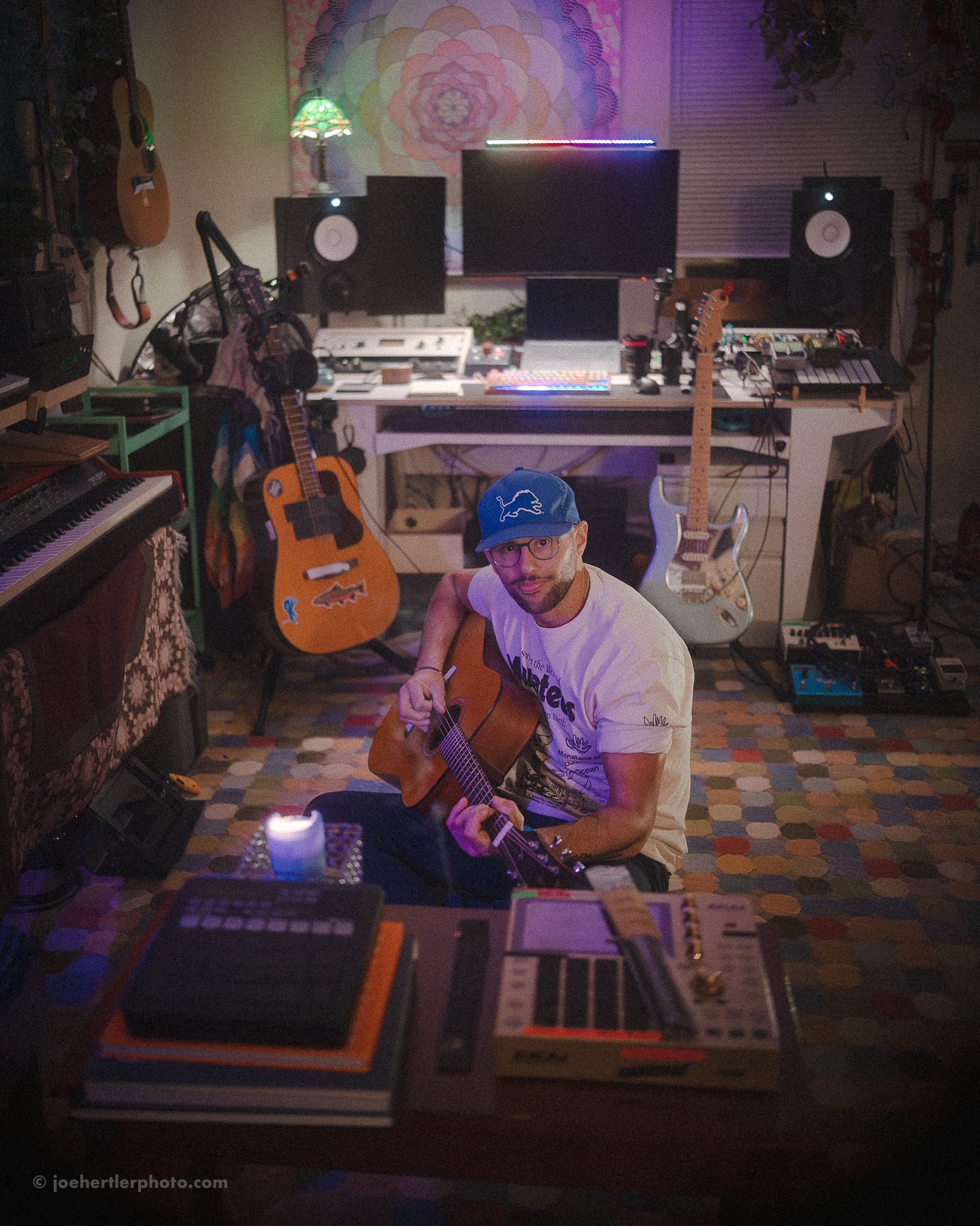 A man wearing a blue cap, glasses, and a white T-shirt is sitting on the floor and playing an acoustic guitar in a music studio. The studio has multiple guitars, keyboards, a microphone, computer monitors, and studio equipment. The room has colorful 