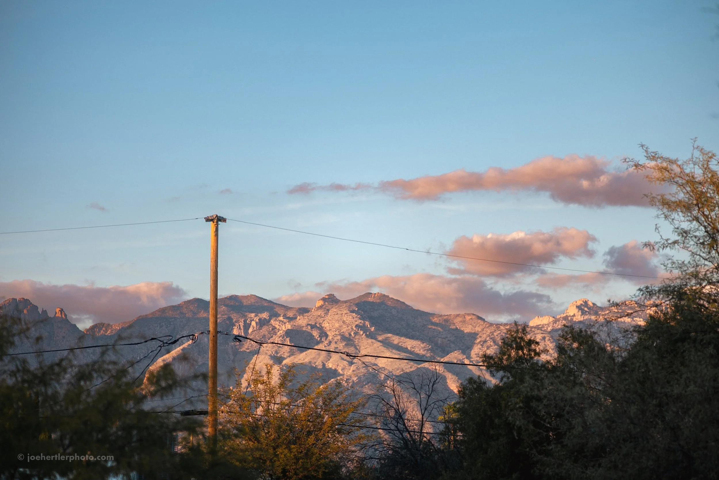 Mountains with rocky peaks illuminated by sunset, partly cloudy sky, power pole with wires, trees in foreground.