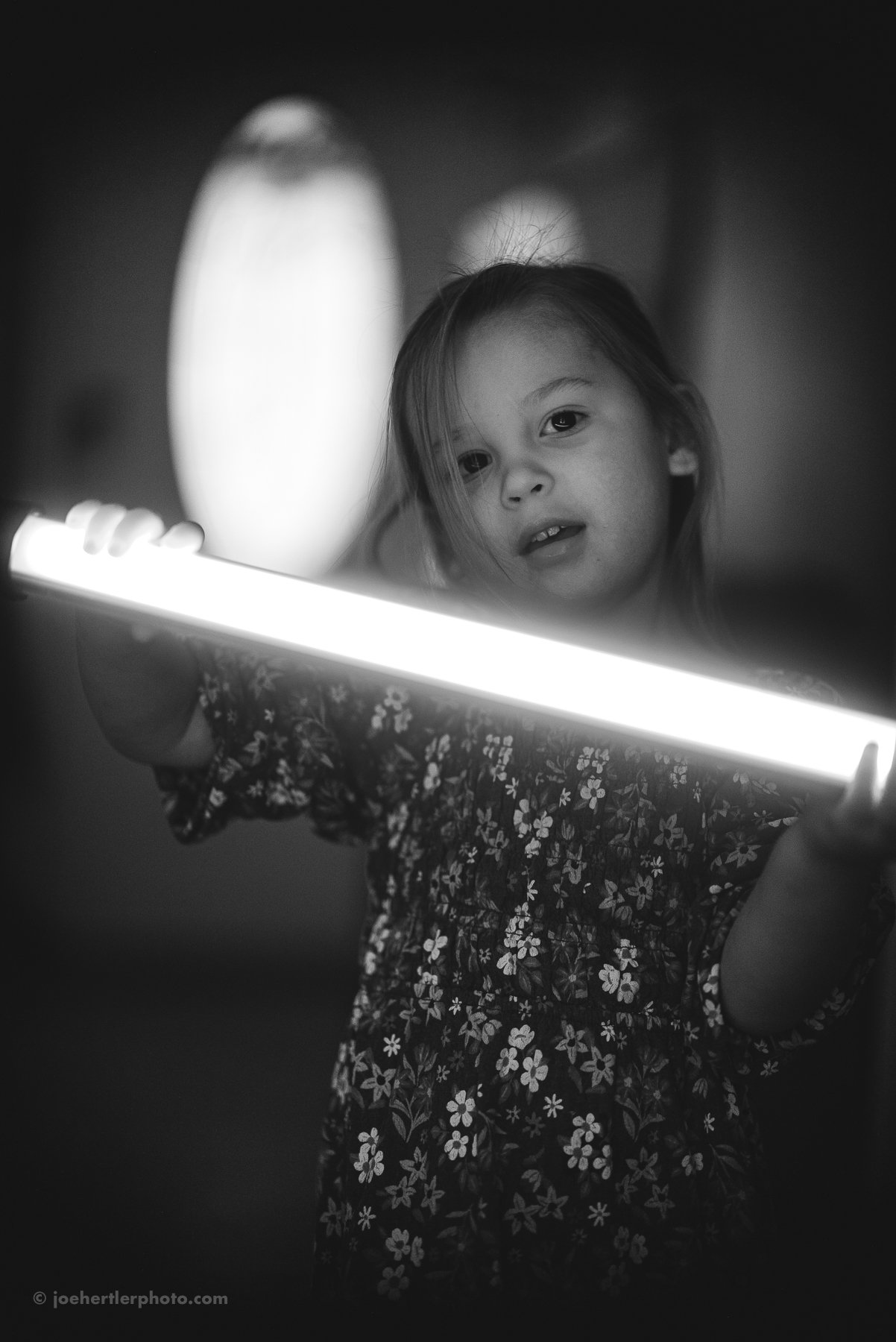 A young girl with light-colored hair, wearing a floral dress, is holding a bright light tube in a dark environment. Her face is illuminated by the light, and she is looking towards the camera with a curious expression.