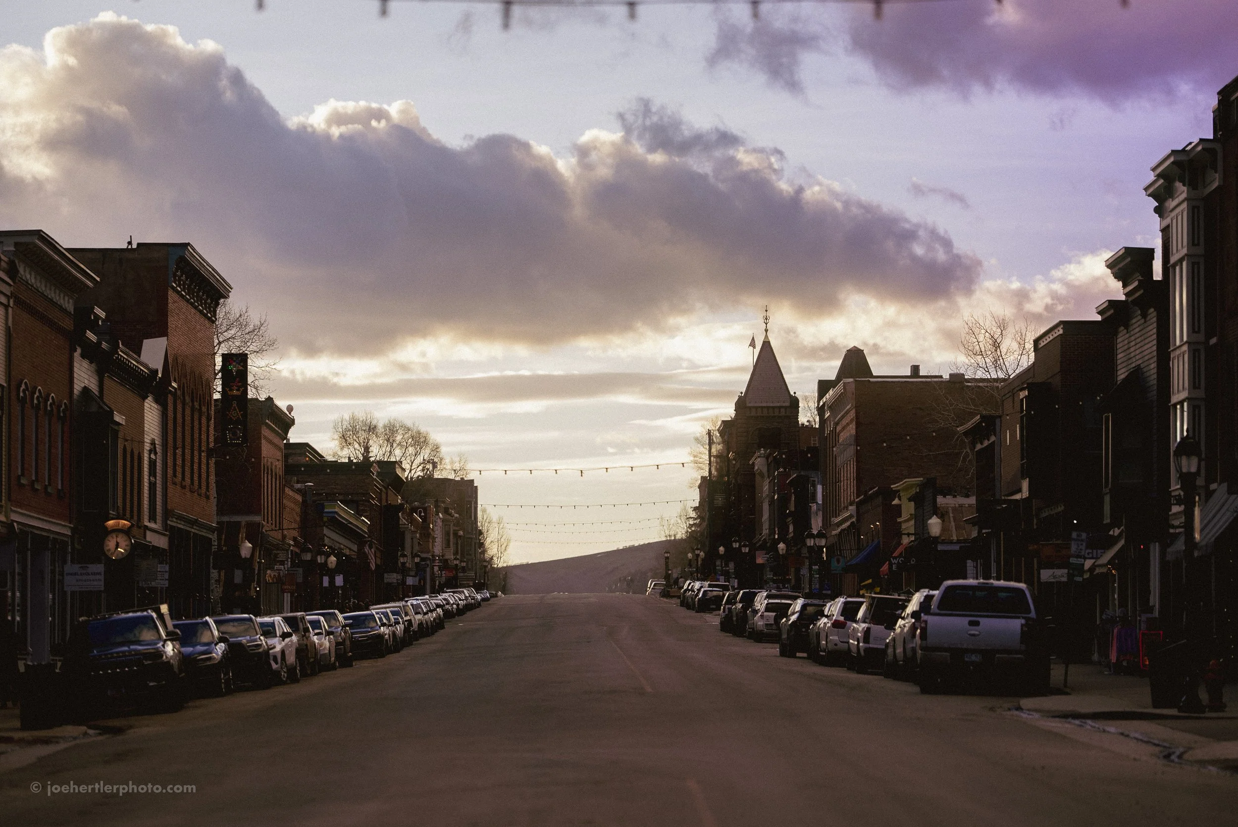 A small town street at sunset with parked cars along both sides and historic buildings featuring brick facades. The sky is cloudy with the sun setting in the distance, creating a moody atmosphere.