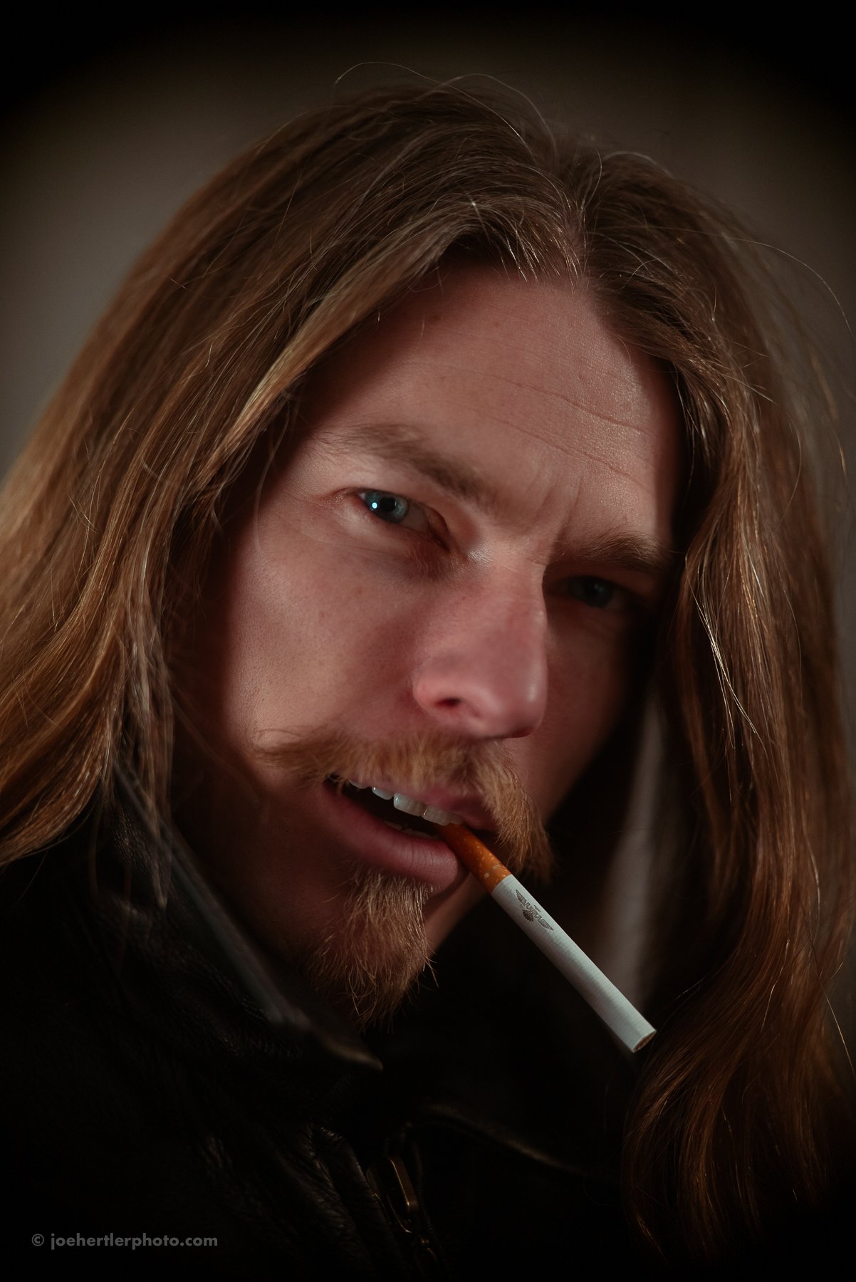 Close-up of a man with long brown hair and a beard, holding a cigarette in his mouth, looking at the camera.