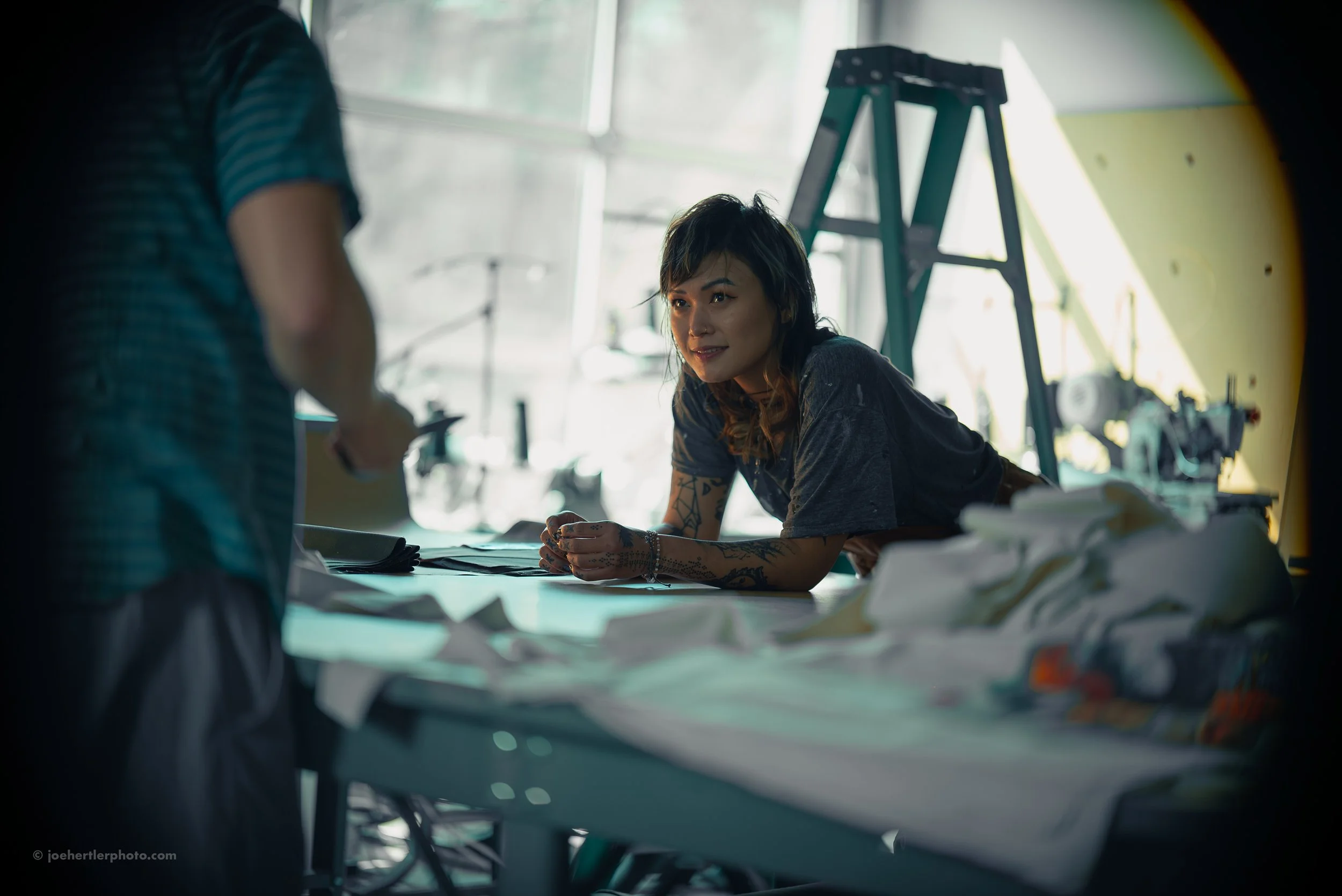 A woman with tattoos and dark hair is sitting at a table with fabric and papers, engaging with a person standing nearby in a workshop or creative studio, with a ladder and crafting equipment in the background.