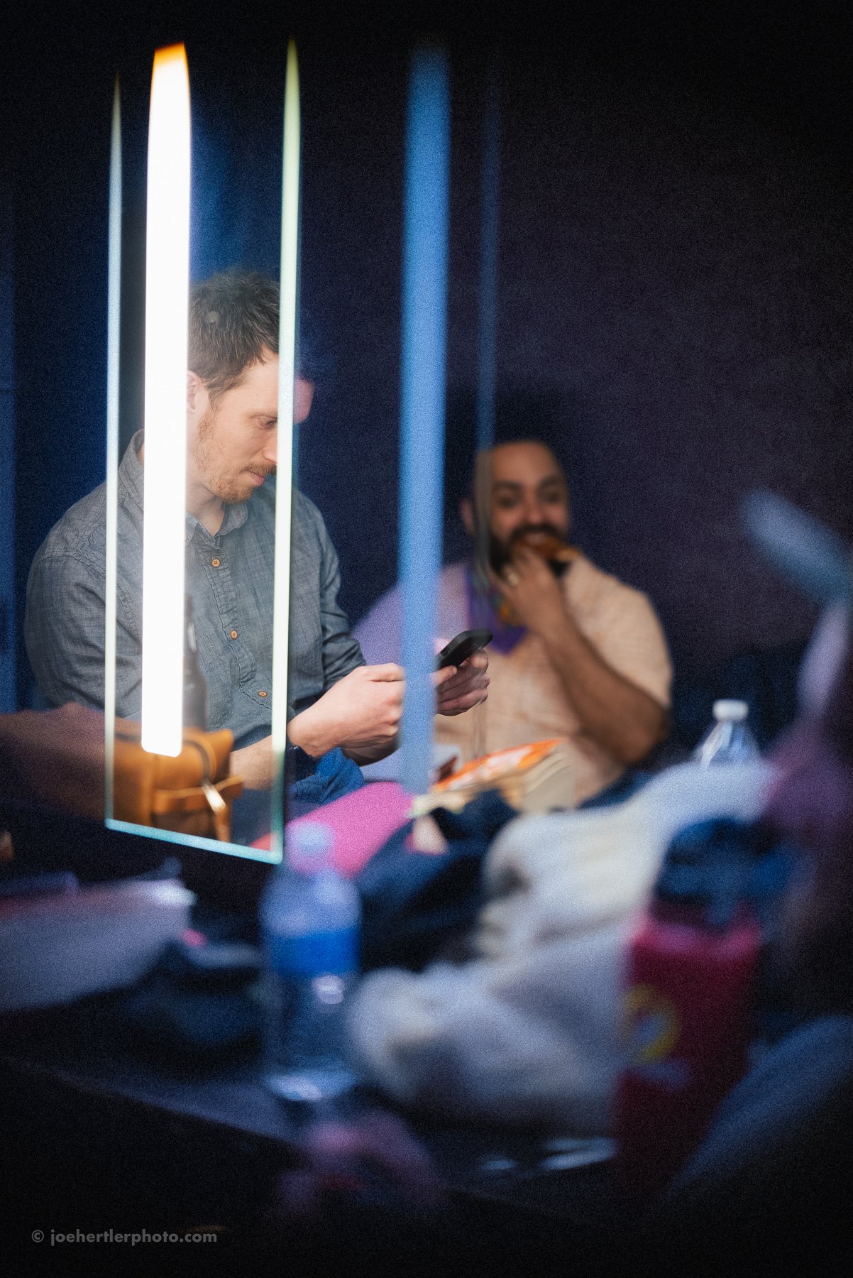 Two men sitting at a table in a dimly lit room, one looking at his phone and the other smiling and touching his beard, with various items and water bottles on the table.