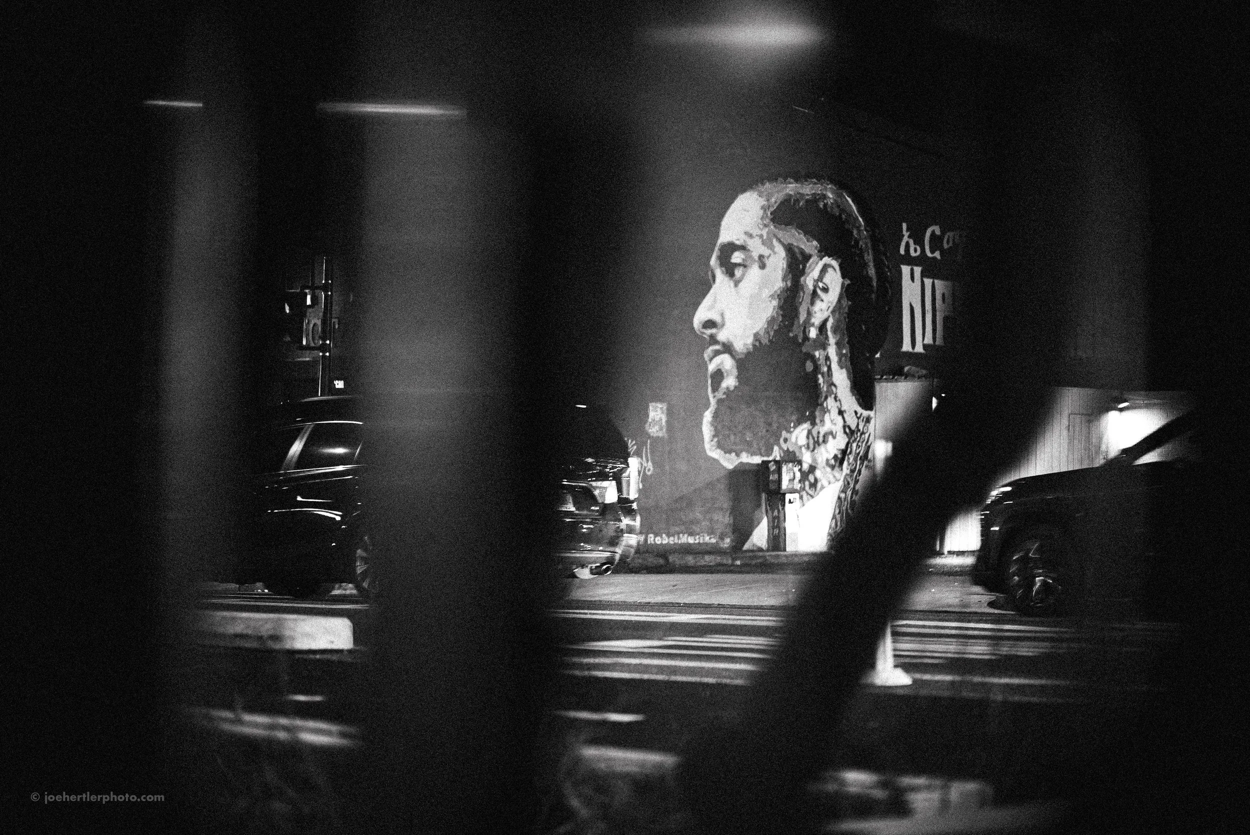 Black and white photo of a street scene at night, showing a large mural of a man with facial tattoos and long hair on a building wall. Several parked cars are visible in front, partially obscured by dark foreground elements.