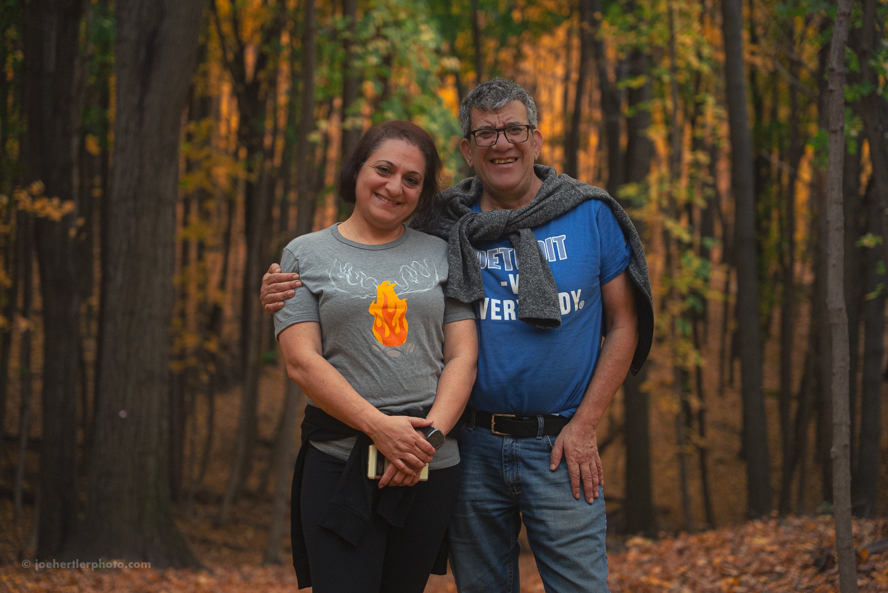 A smiling woman and man standing together in a forest during fall, with colorful autumn leaves on the ground and trees around them. The woman is wearing a gray t-shirt with a flame design, and the man is wearing a blue t-shirt with text, with a gray 
