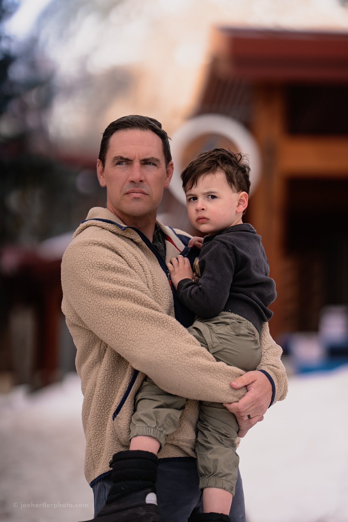 A man holding a young boy outdoors on a winter day with snow on the ground and a blurred background of a building.