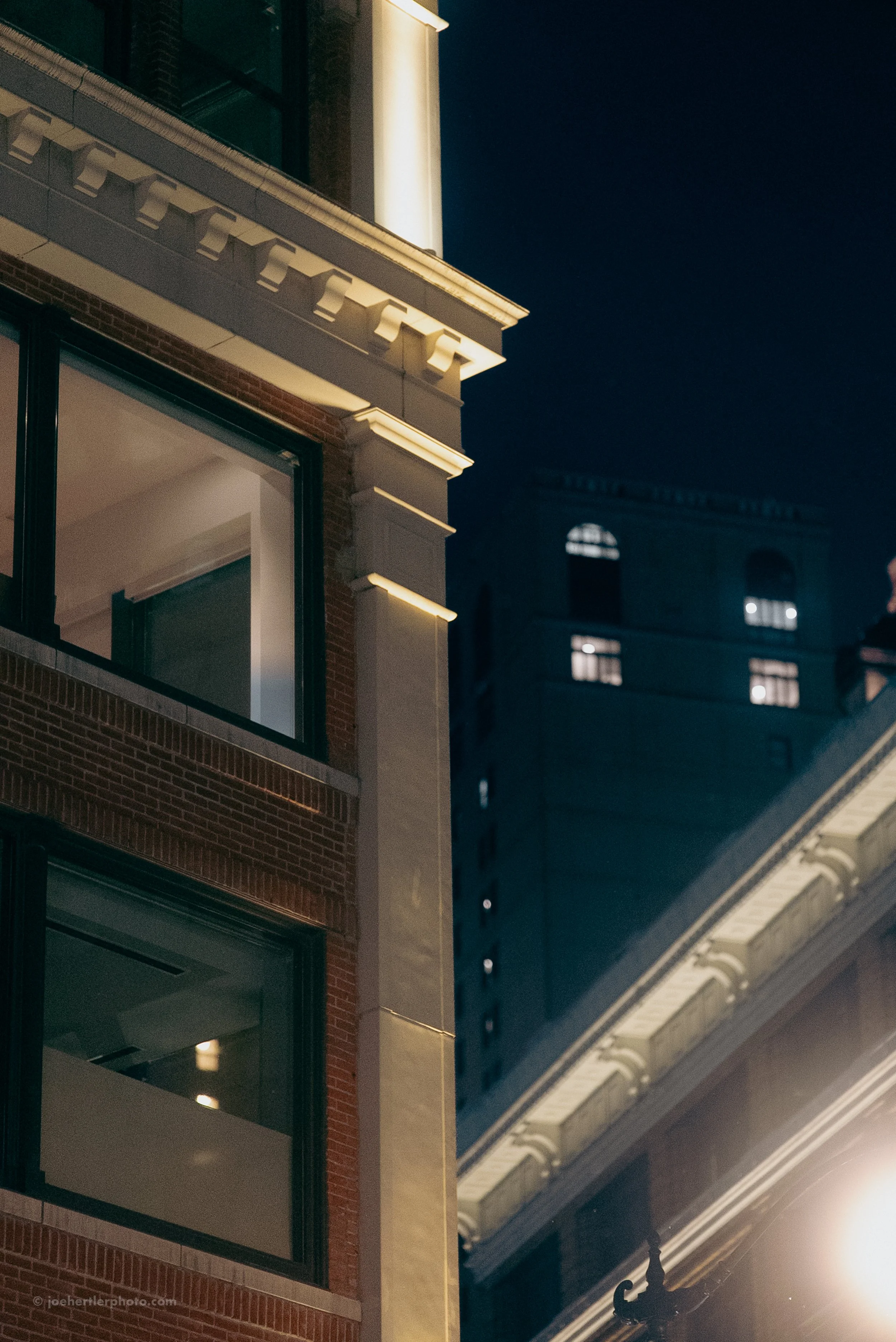 Nighttime view of a multi-story building's corner with illuminated architectural details, and a tall dark building with lit windows in the background.