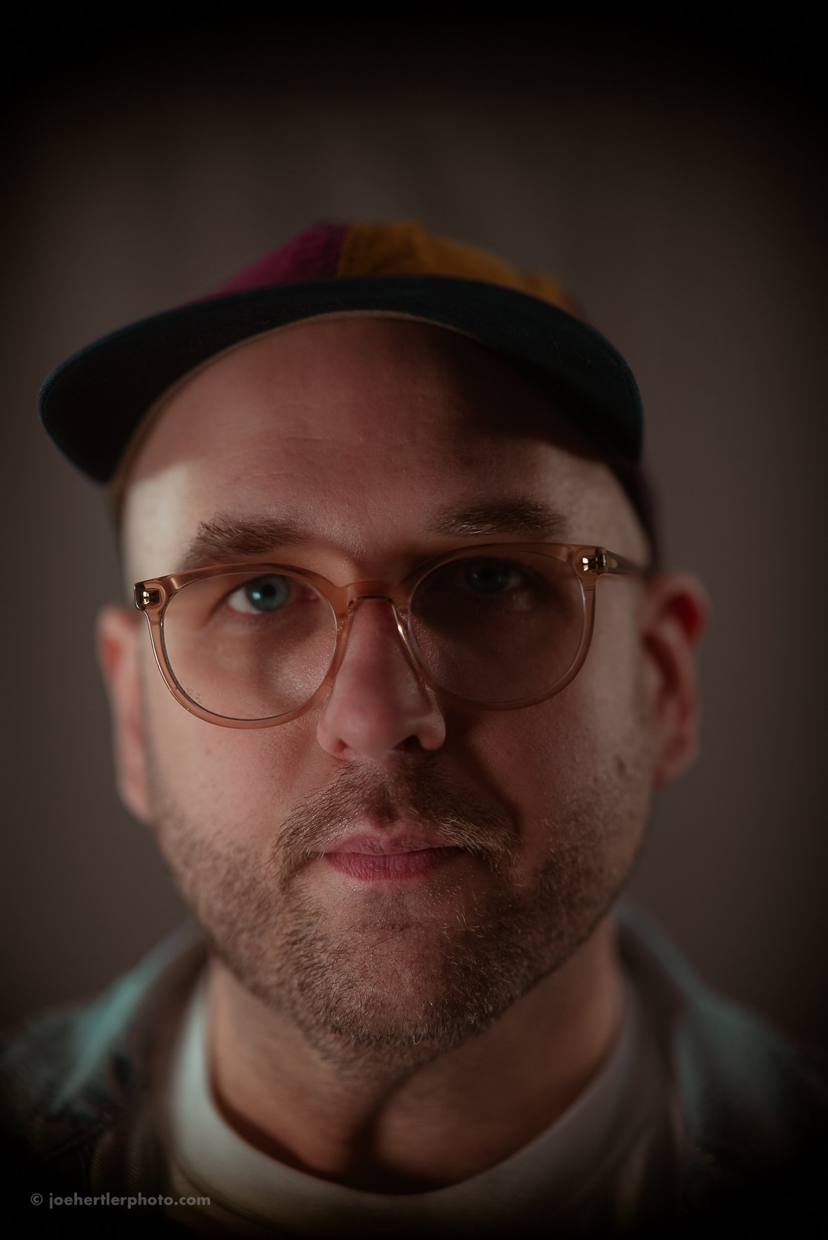 Close-up portrait of a man wearing glasses and a cap, with a neutral expression, blurred background.
