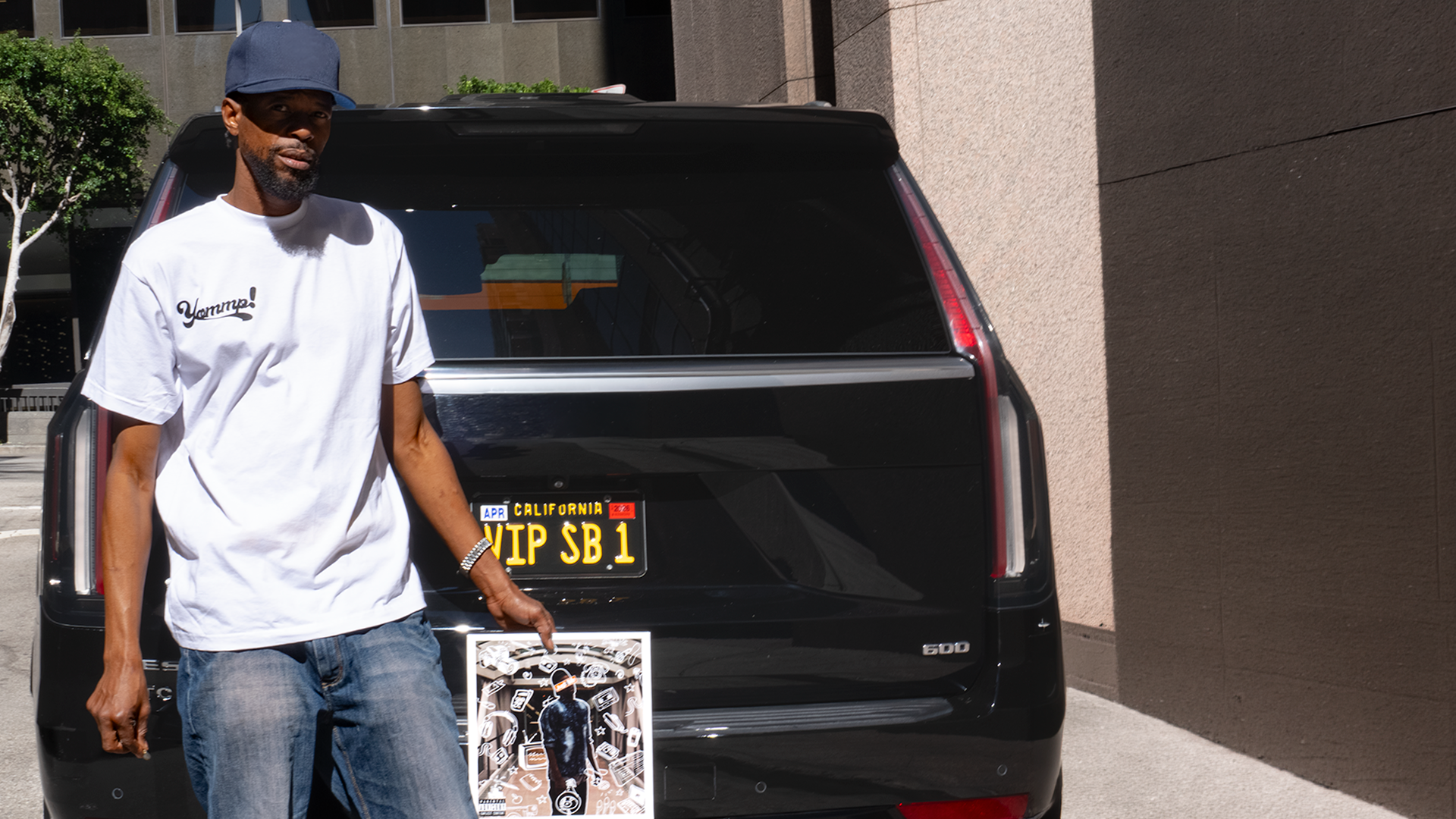A man in a white t-shirt and blue cap standing next to a black SUV with California license plates, holding a poster.