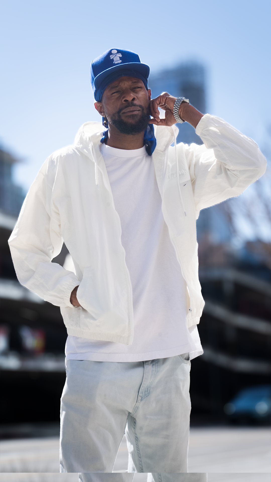 A man standing outdoors on a sunny day, wearing a white jacket, white T-shirt, light jeans, a blue cap, and a watch, with urban buildings and a blurred cityscape in the background.