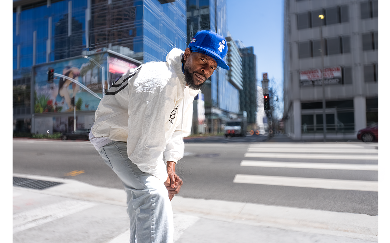 A man with a beard crouching on the sidewalk at an intersection in an urban area, wearing a white jacket, light jeans, and a blue cap, looking at the camera with an intense expression.
