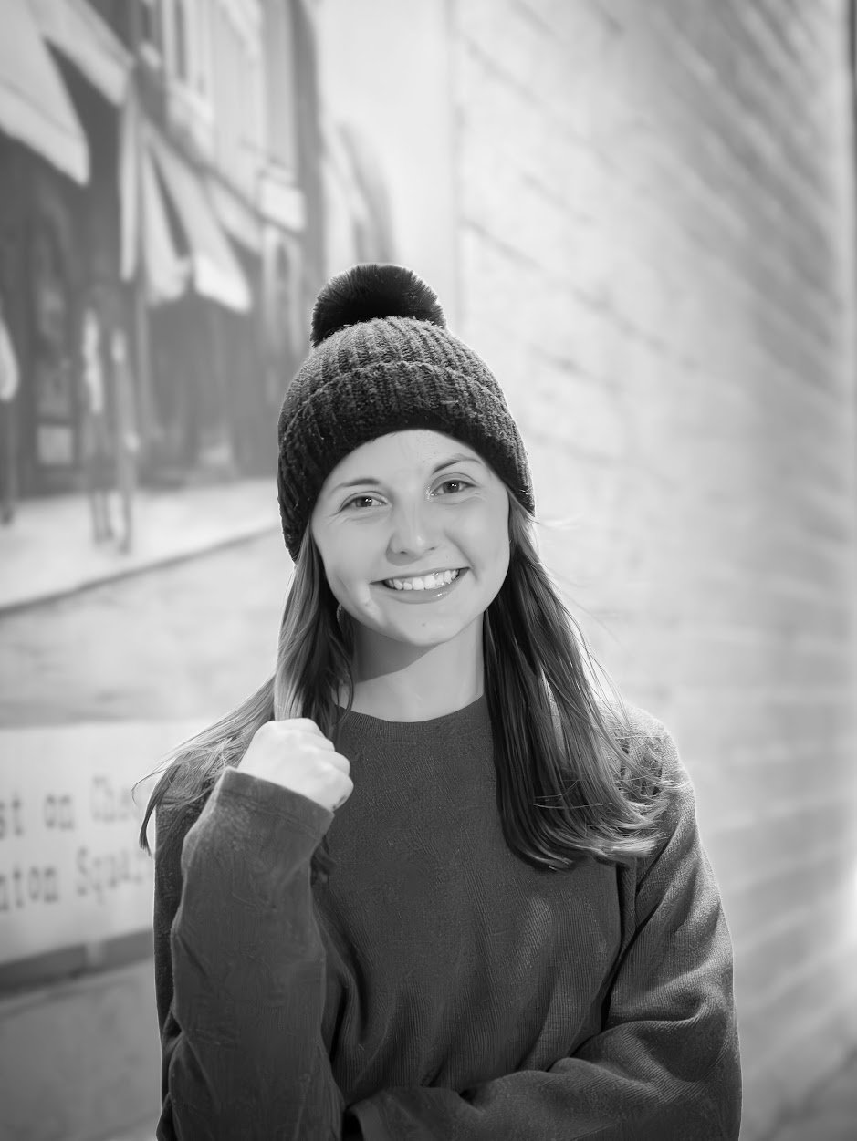 A smiling young woman wearing a winter beanie and sweater, posing with her fist clenched in a gesture of confidence indoors.