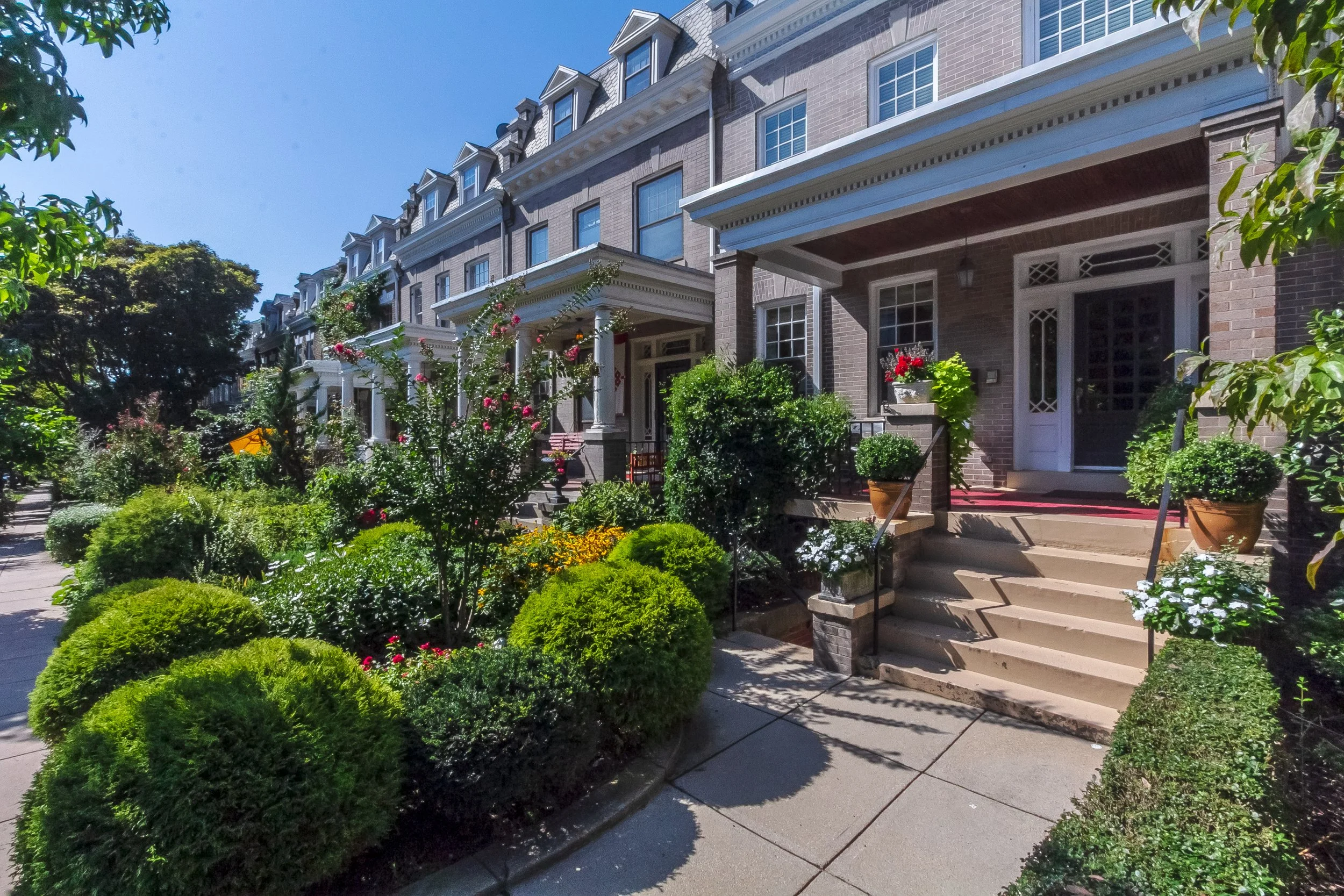 Row of brick townhouses with front porches and stairs, surrounded by well-maintained garden with bushes, flowering plants, and potted plants. Clear, sunny day.