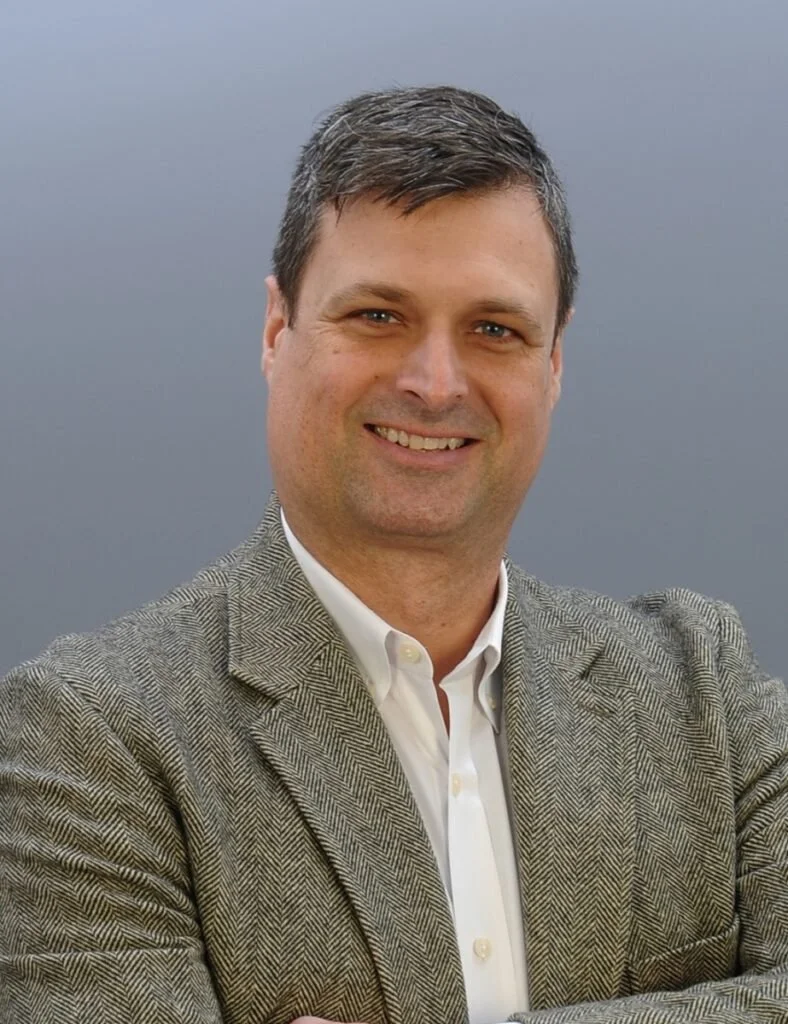 Drew Christian in a gray herringbone blazer and white shirt, posing in front of a plain gray background.