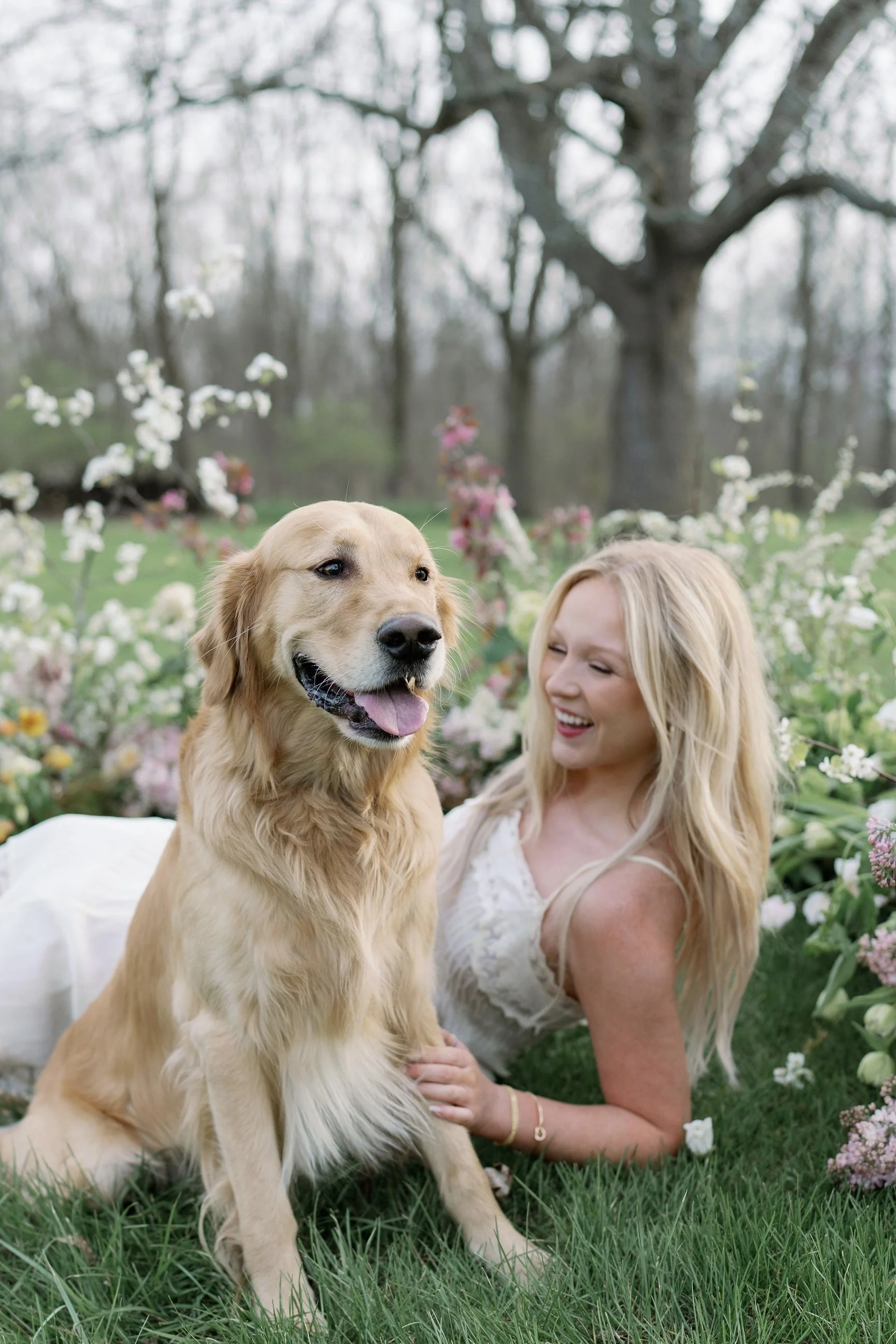 A woman with blonde hair, wearing a white dress, lying on grass surrounded by flowers, smiling and looking at a golden retriever dog. The dog is sitting in front of her, panting with its tongue out, outdoors in a garden with trees.