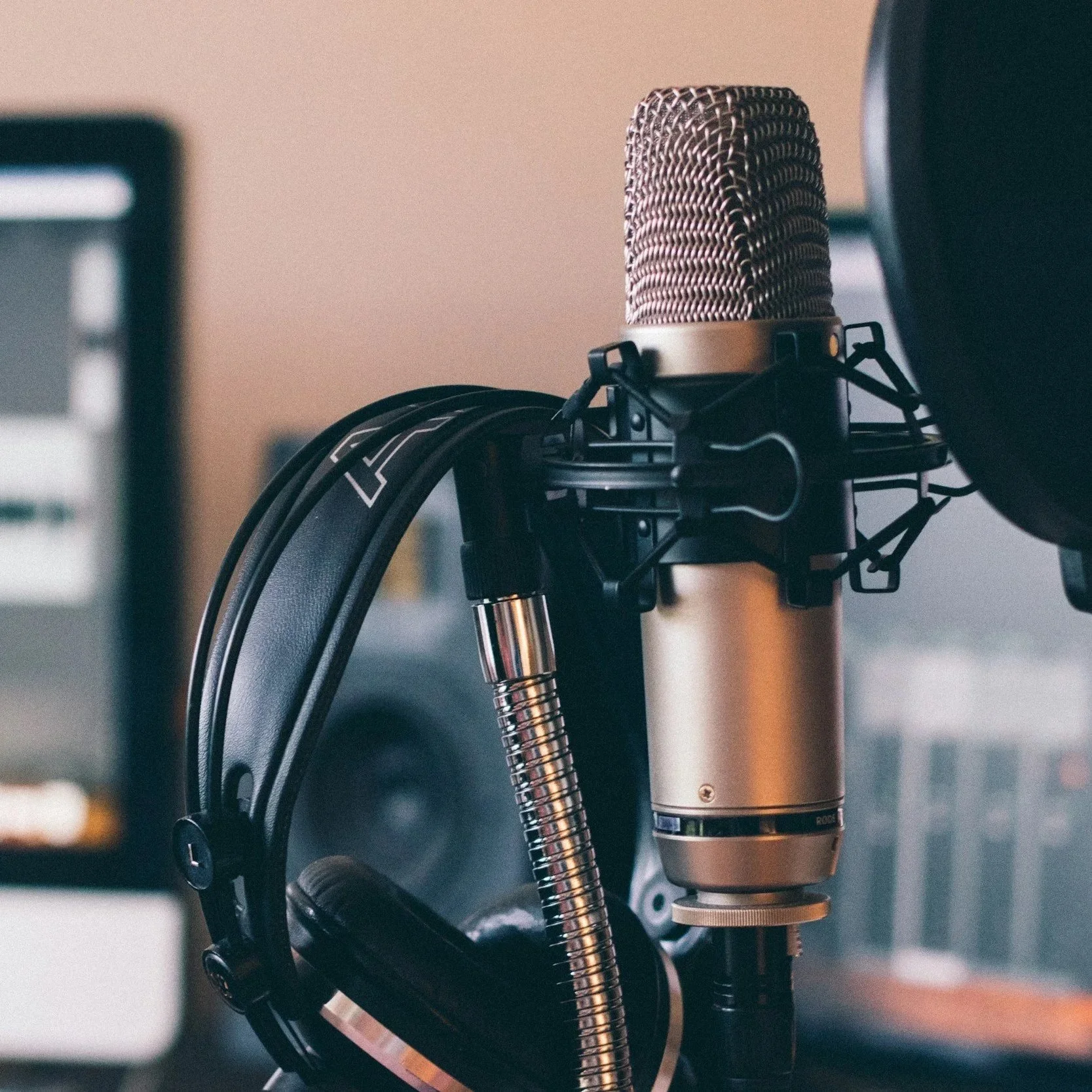 A professional studio microphone mounted on a pop filter, with headphones hanging nearby and a computer monitor in the background.