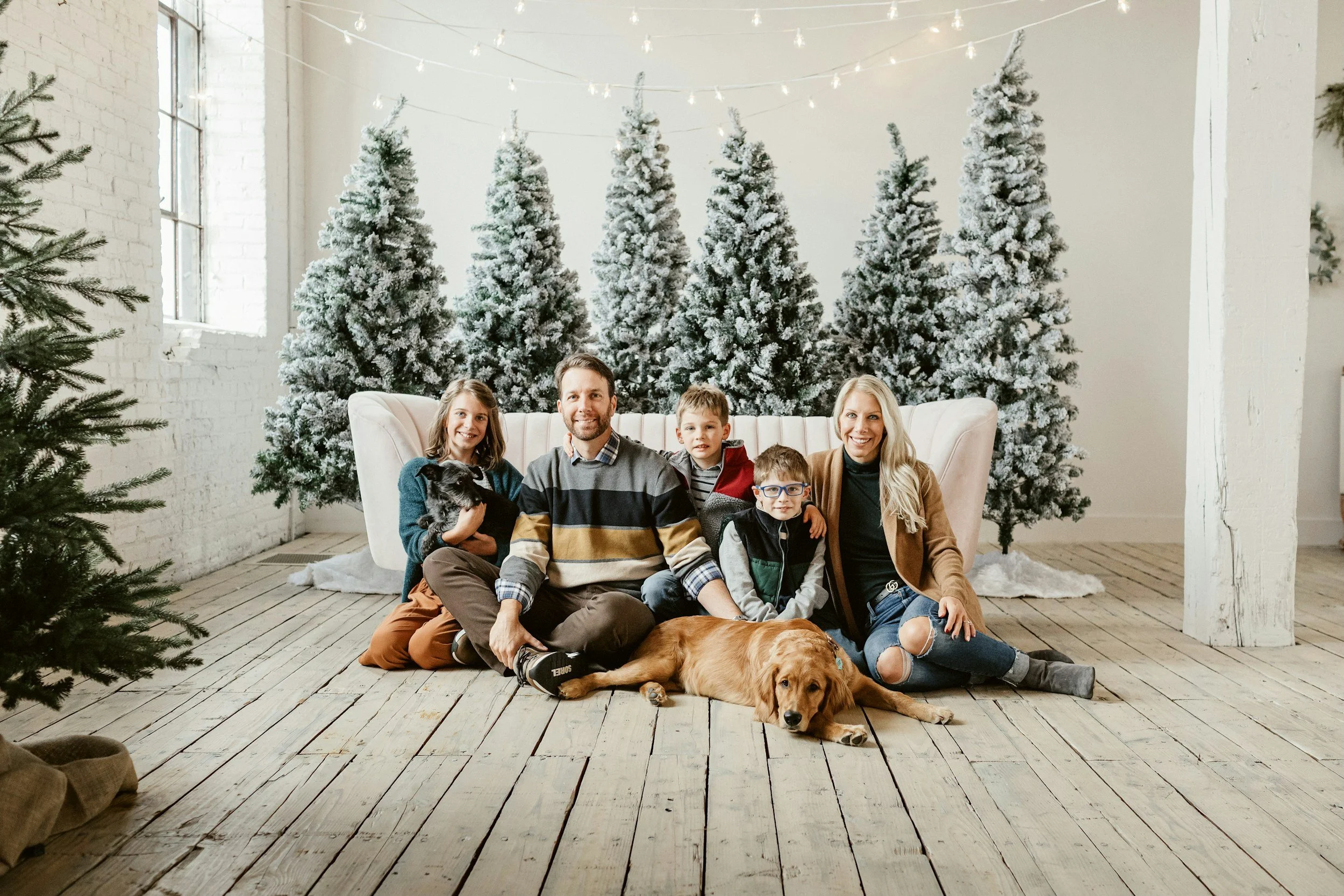 Family of six, including two children, sitting on the wooden floor in front of a white sofa, with three snow-dusted Christmas trees and string lights in the background, and a dog lying on the floor in a festive indoor setting.
