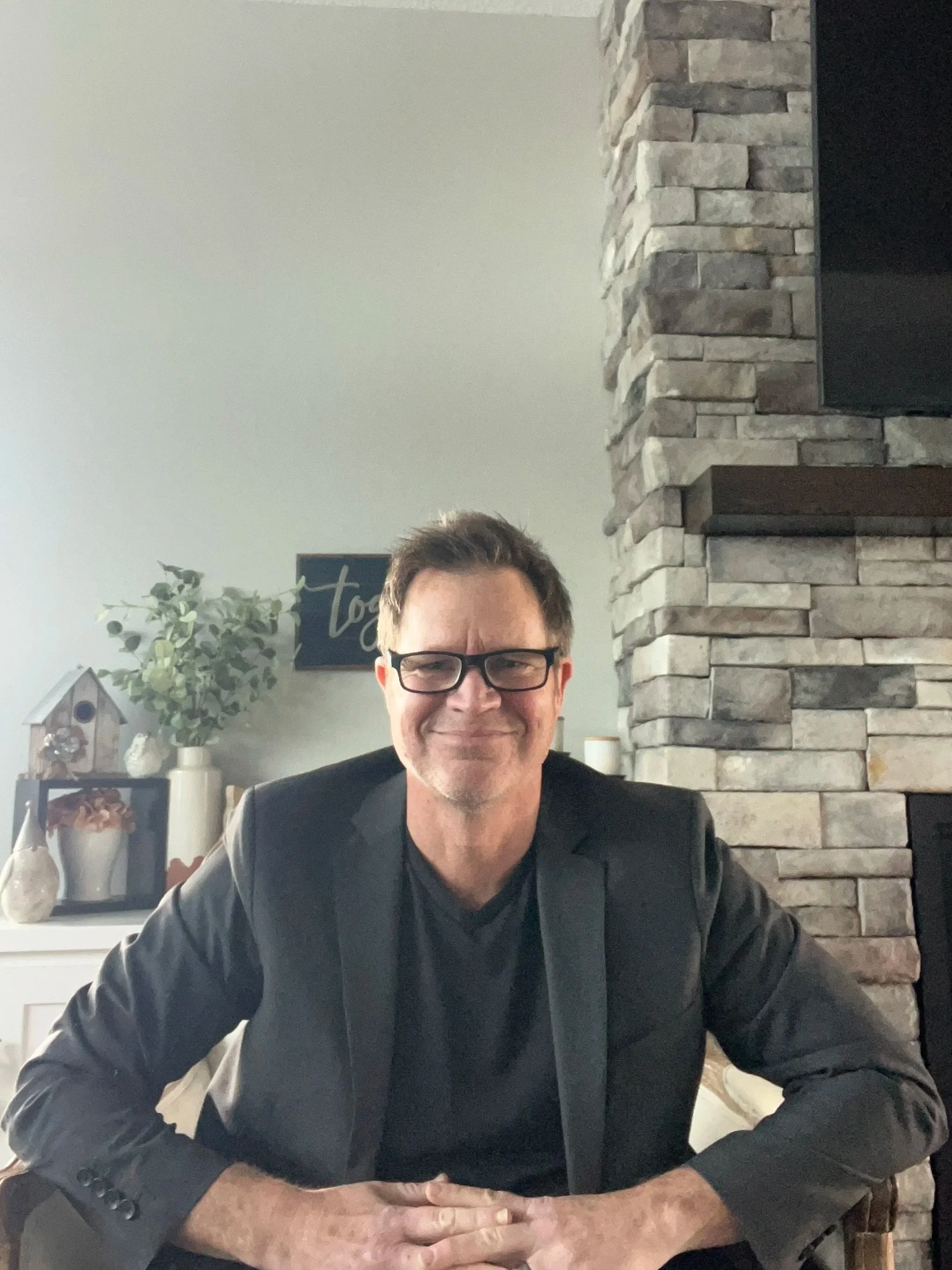 A man with glasses and dark hair, smiling, sitting at a table in a living room with a stone fireplace and decorative items in the background.