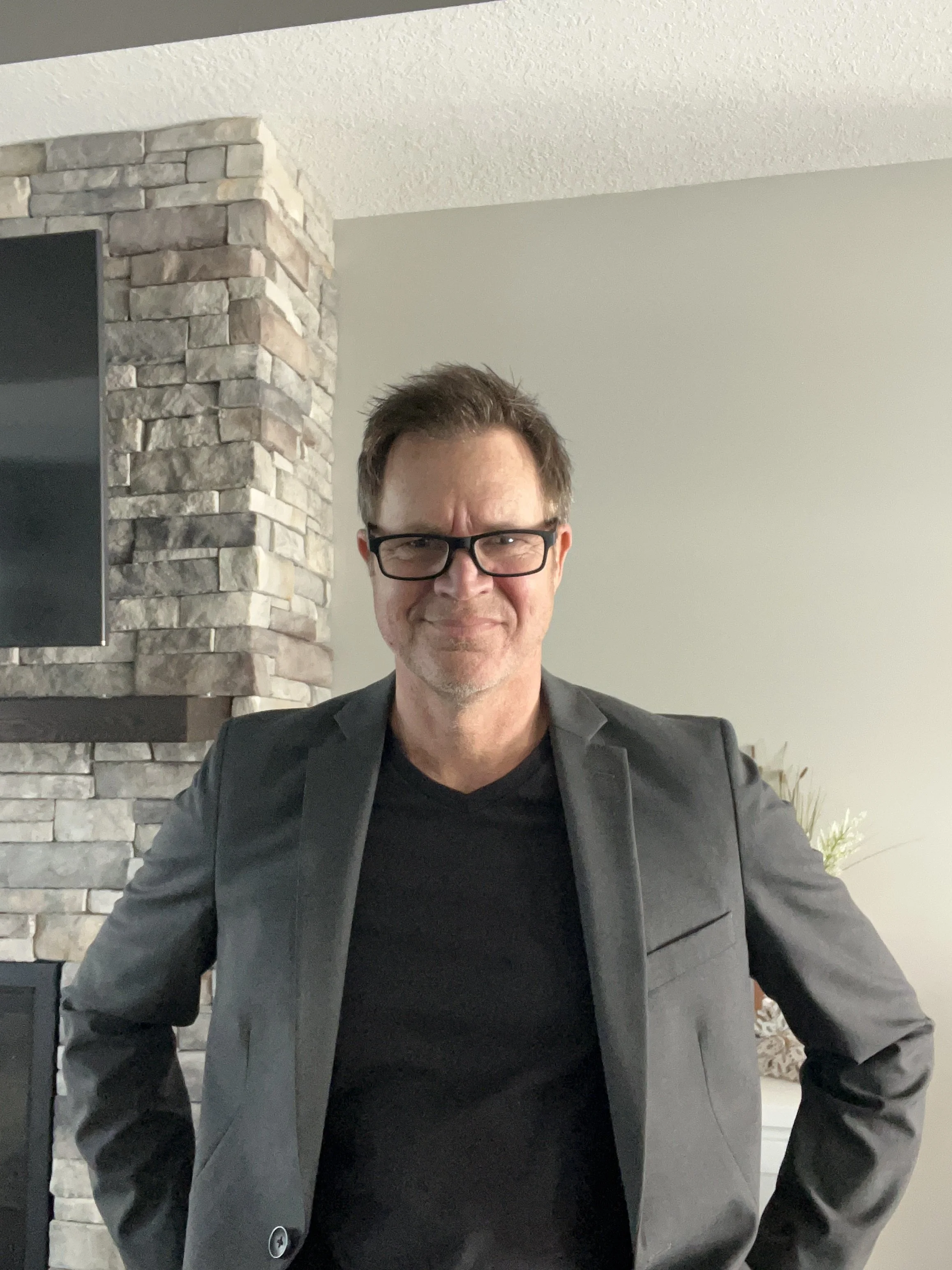 A man with glasses, brown hair, and a light beard, wearing a black shirt and a gray blazer, standing indoors in front of a stone fireplace and a light-colored wall.