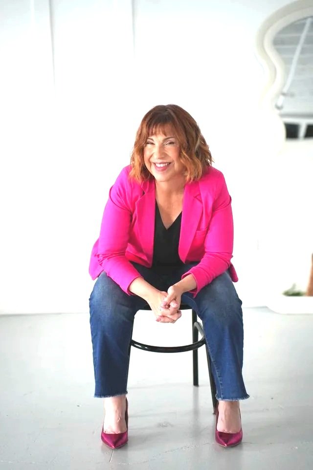 Woman sitting on a chair laughing, wearing a bright pink blazer, black top, blue jeans, and pink high heels, in a brightly lit room.