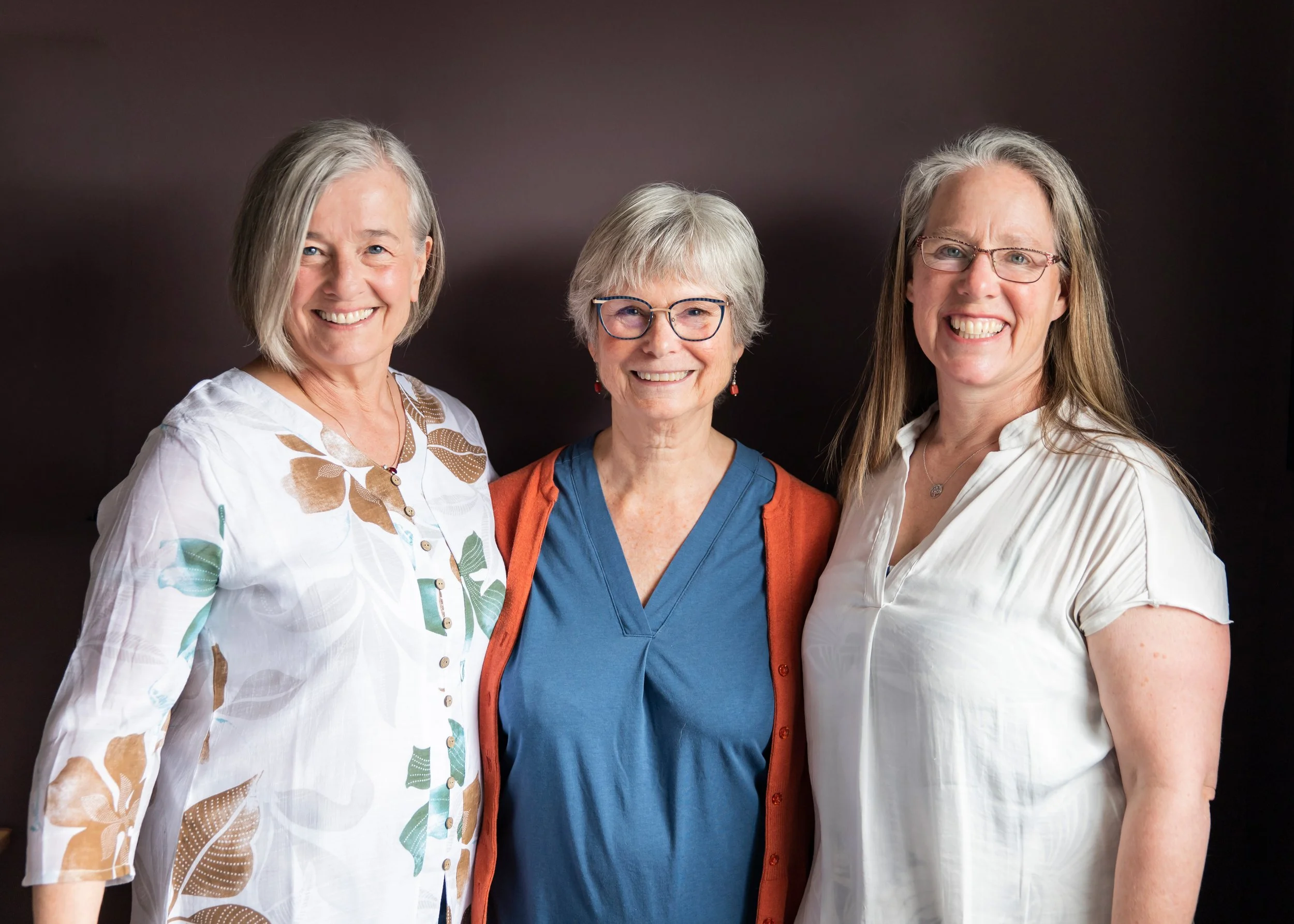 Three smiling women standing together against a black background, dressed in casual clothes, with the woman in the middle wearing glasses.
