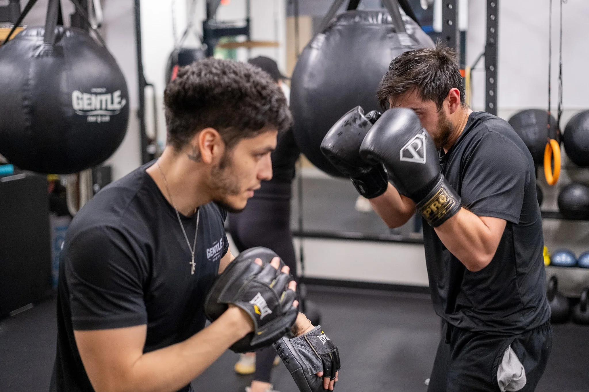 Two men training with boxing gloves in a gym. One man is kneeling, and the other man is standing, holding his gloves up in a fighting stance.