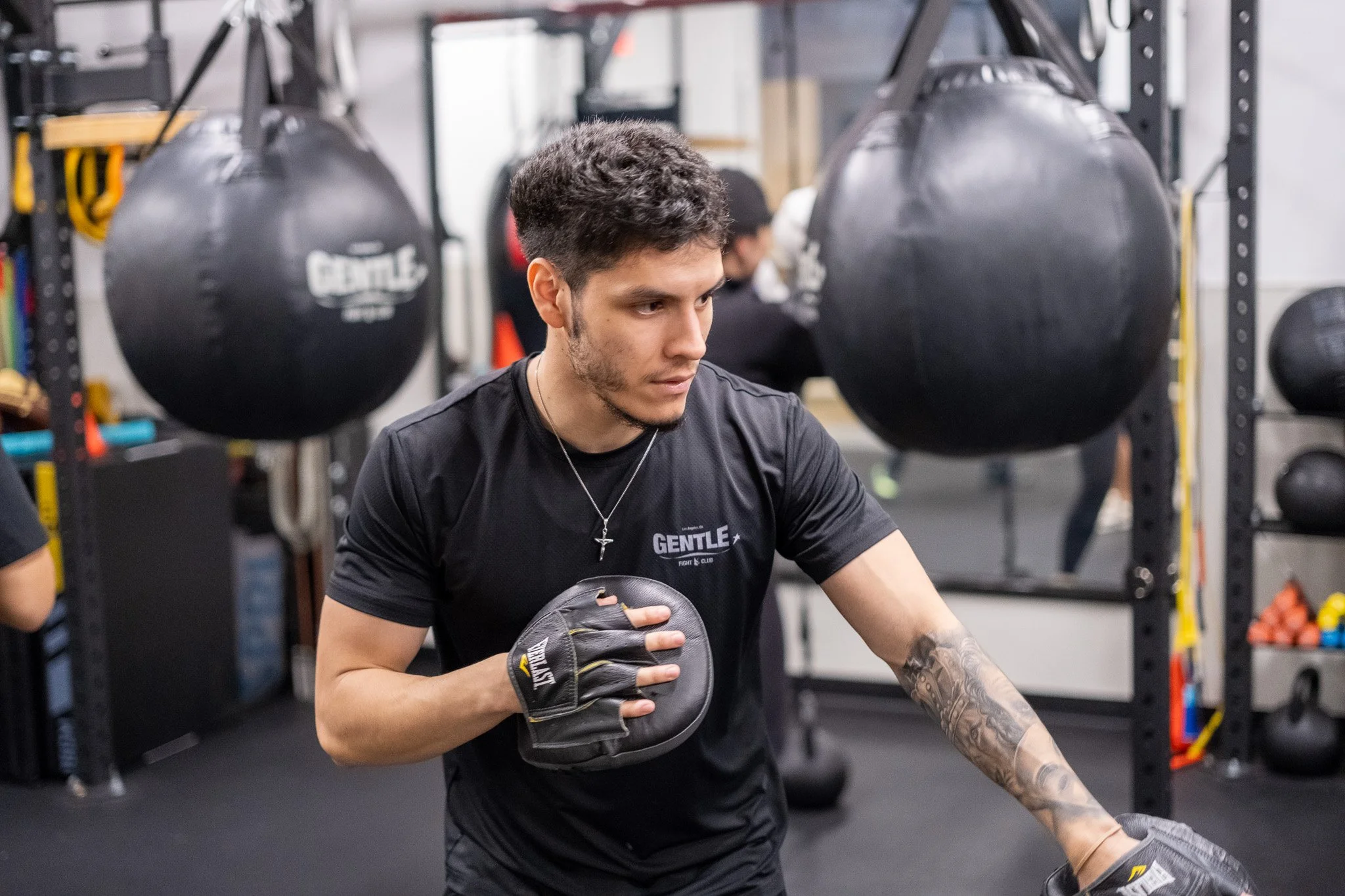 A young man in a gym practicing boxing, holding a focus mitt and wearing a black T-shirt and boxing glove, with boxing bags hanging in the background.