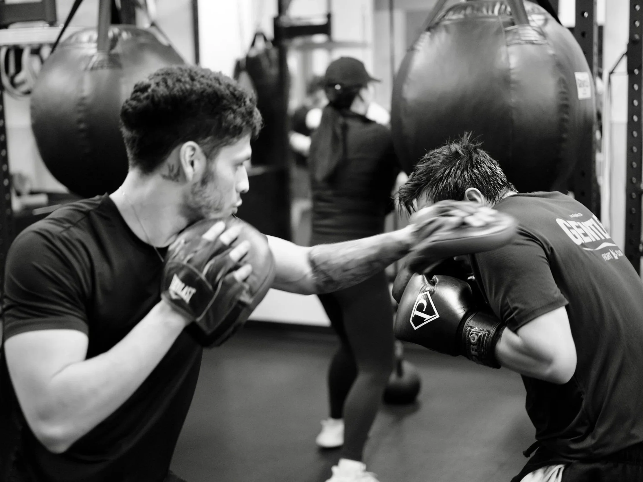 Two young men with athletic physiques engaged in arm wrestling in a gym setting, with other people and training equipment visible in the background, captured in black and white.