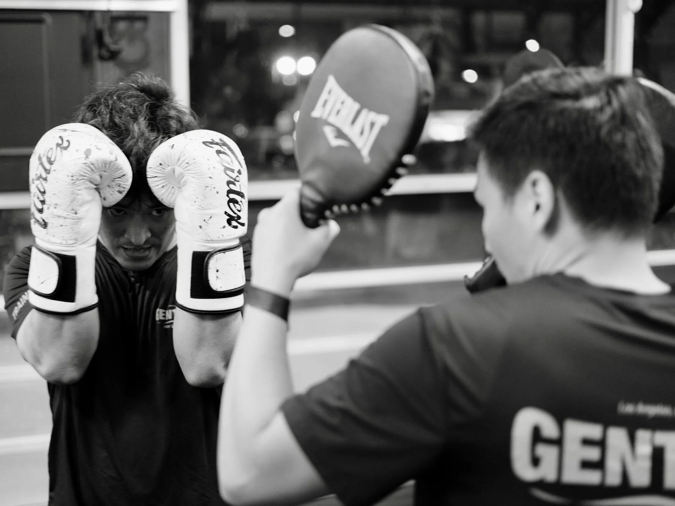 A boxer practicing in a gym, wearing boxing gloves, and staying focused with his opponent or coach in front of him. The background shows part of a boxing ring and some gym equipment.