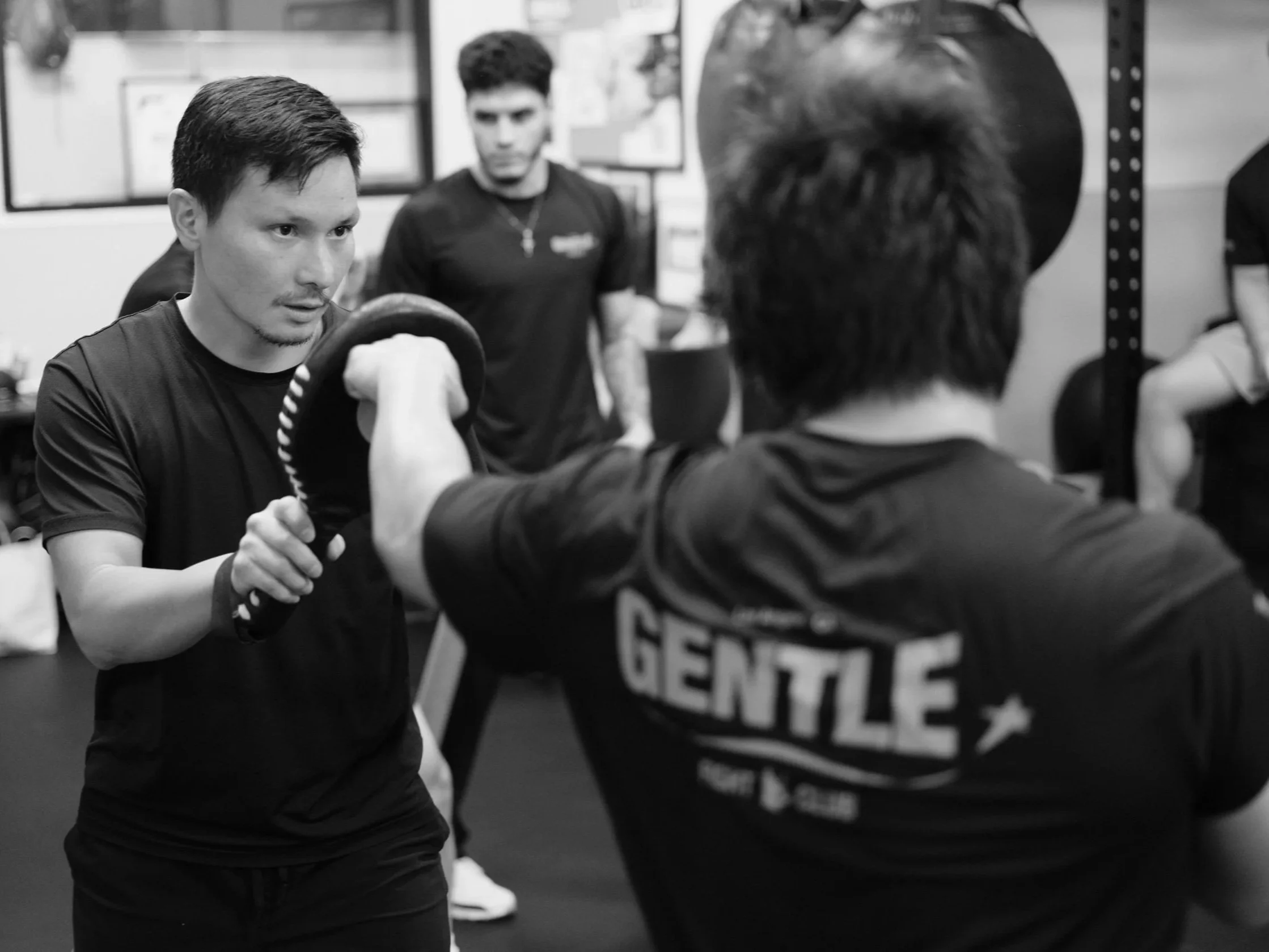 Two men practicing arm wrestling in a gym with one man focused and the other partially visible, with a woman in the background.